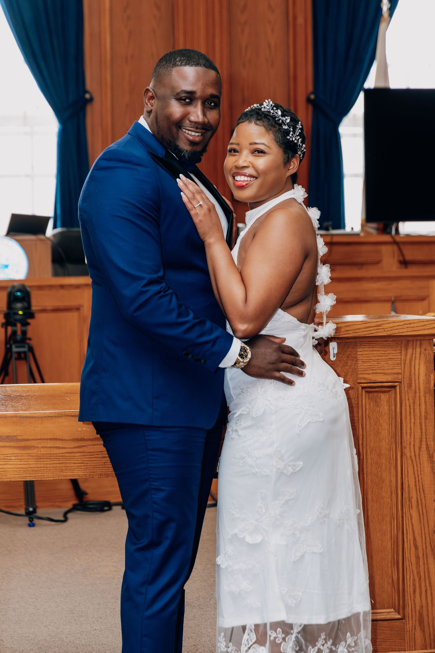 A smiling bride in a white lace gown and tiara poses with her groom in a blue suit inside a church.
