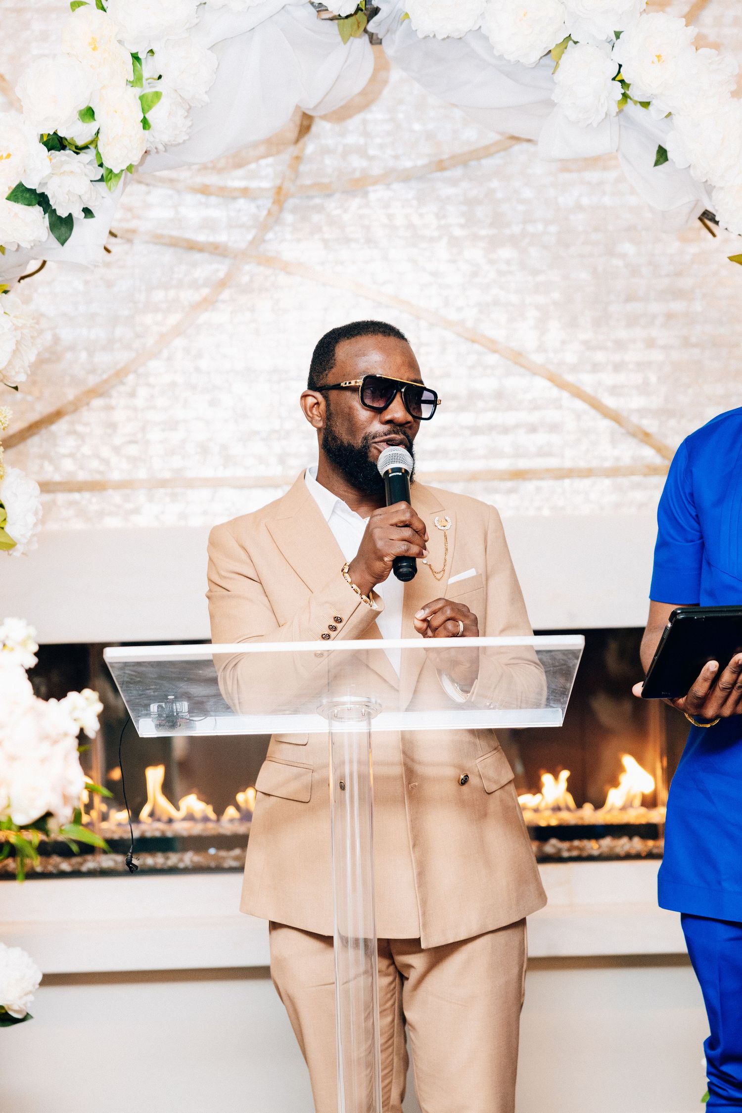 Elegant man in beige suit speaking at wedding ceremony podium with white floral arch and fireplace backdrop.