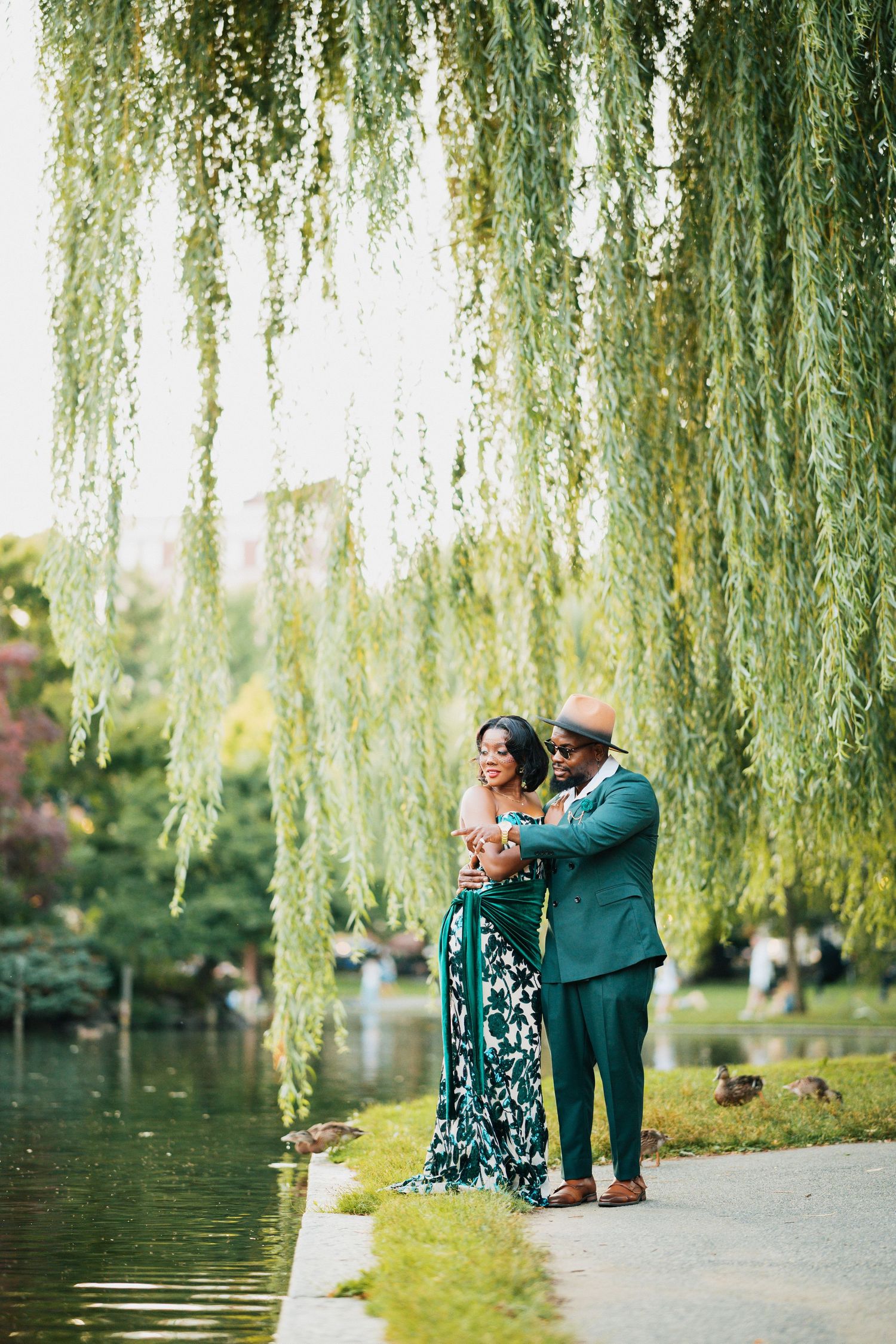 Black couple embracing under weeping willow tree beside tranquil park pond.