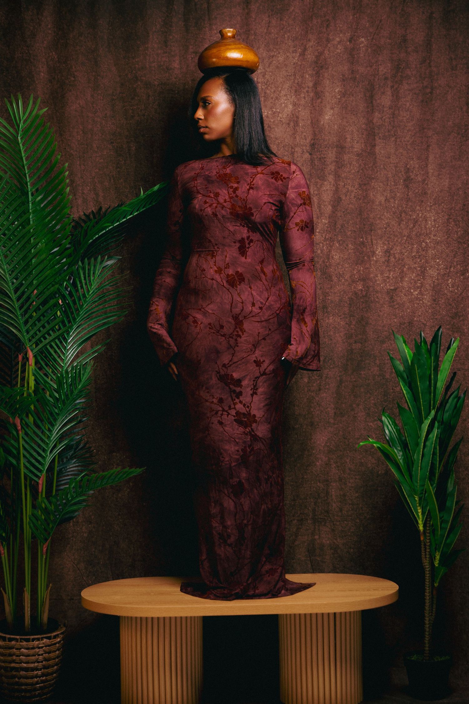 Woman in burgundy lace gown balancing gold vase on head, standing on wooden platform with tropical plants.