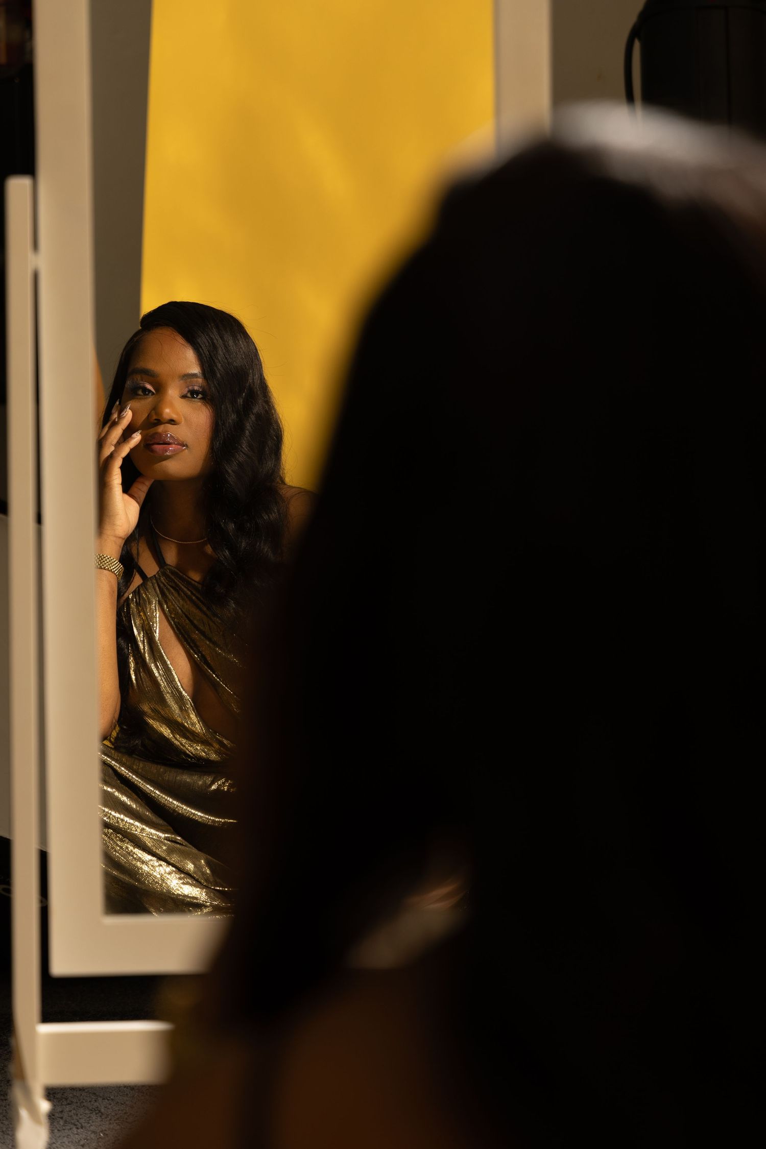 A Black woman in a gold dress applies makeup while seated in front of a mirror with a yellow backdrop.