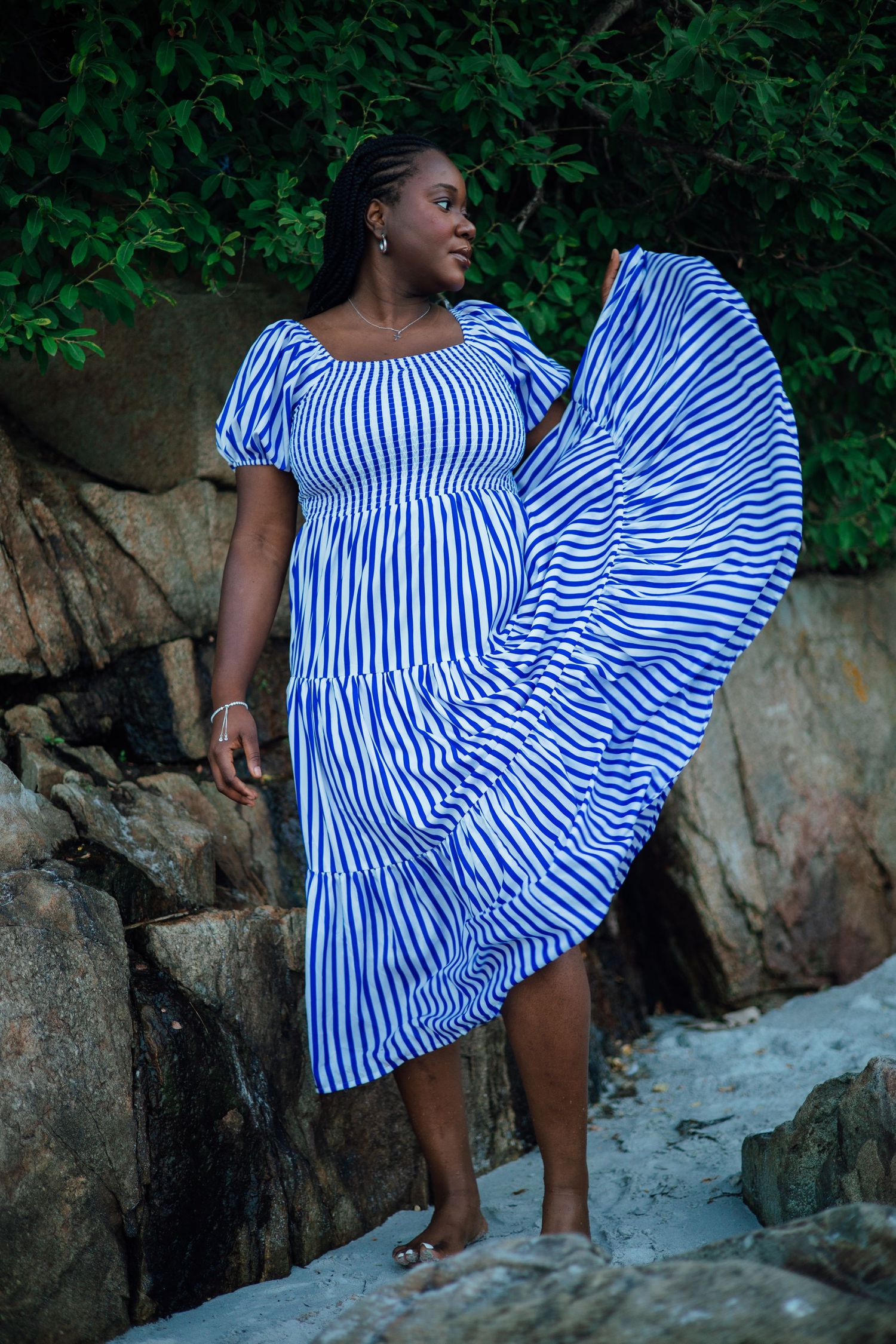 Woman in blue and white striped midi dress posing on rocky beach with wind-blown skirt and green foliage backdrop.