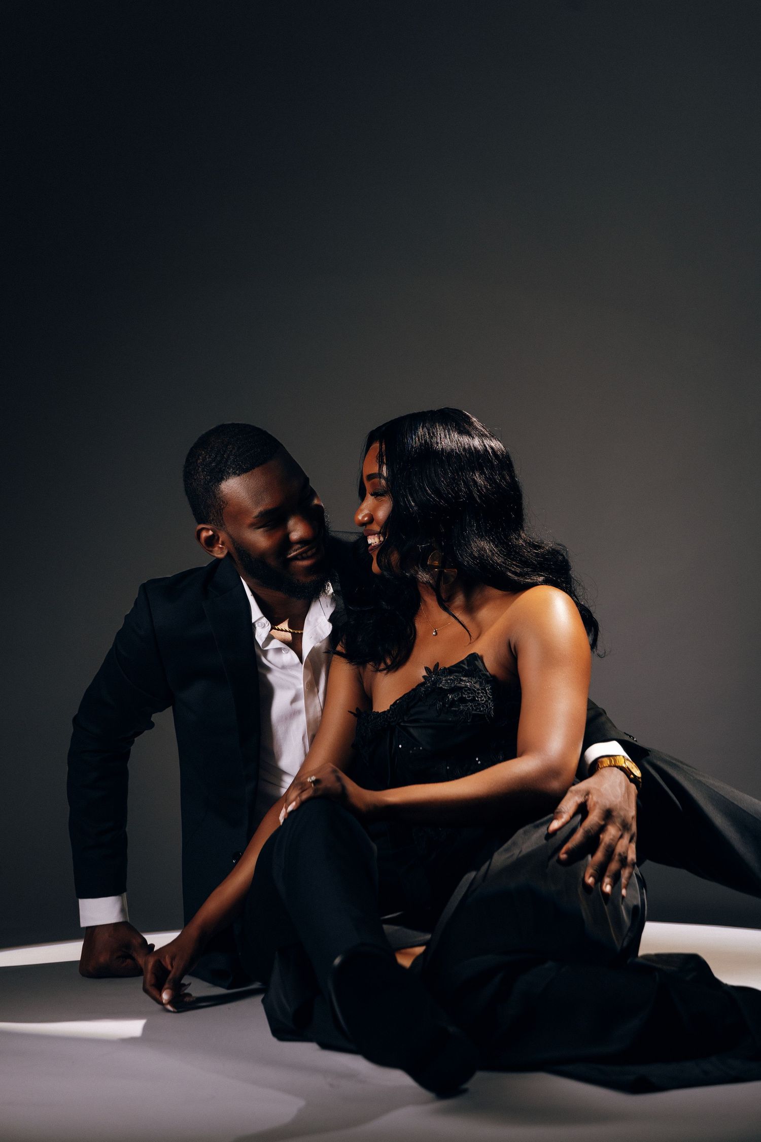 Elegant Black couple in formal attire sitting together on floor, man in suit, woman in strapless gown, studio setting.