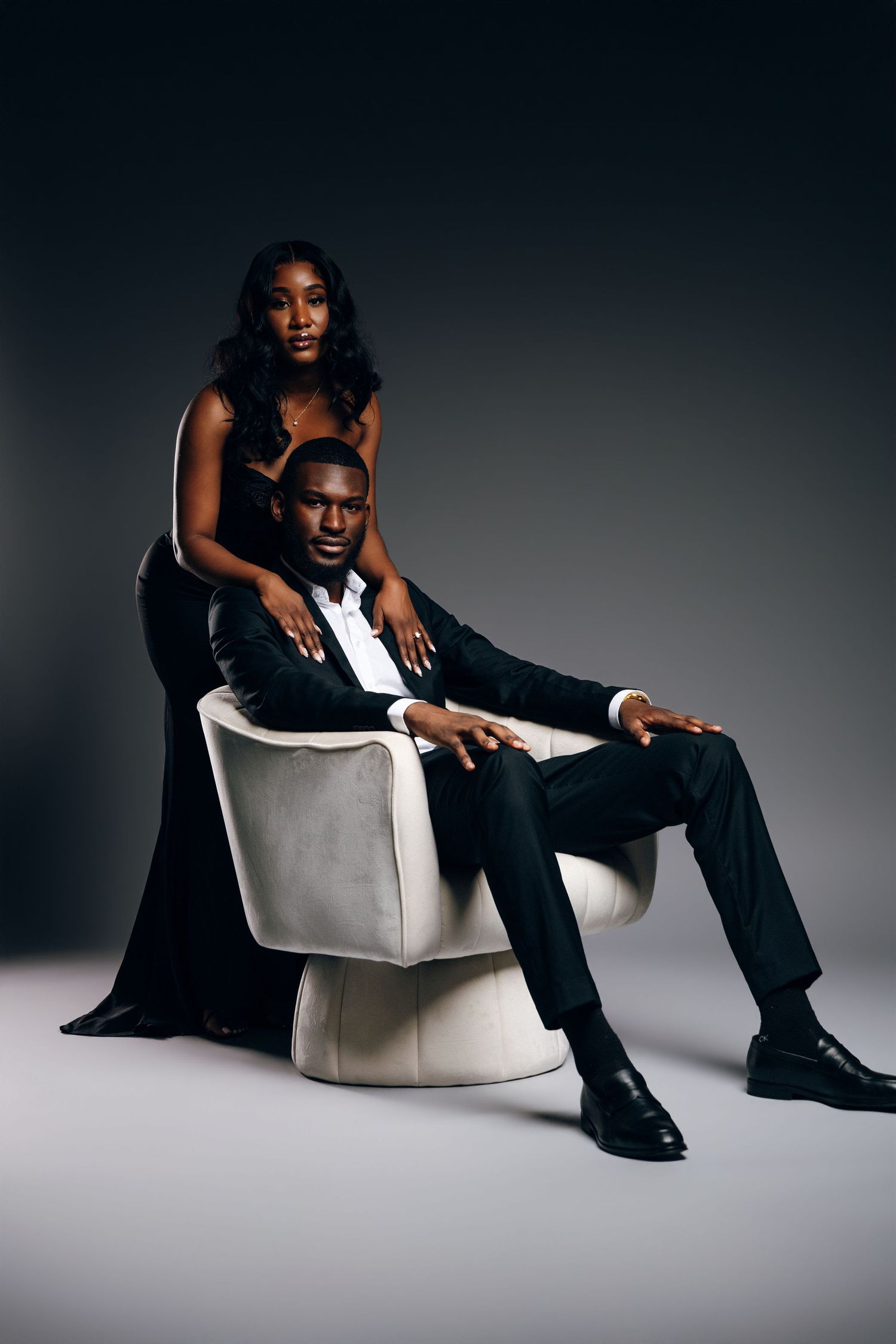Elegant Black couple posing in studio, man seated in white chair wearing suit, woman in black gown standing behind him.