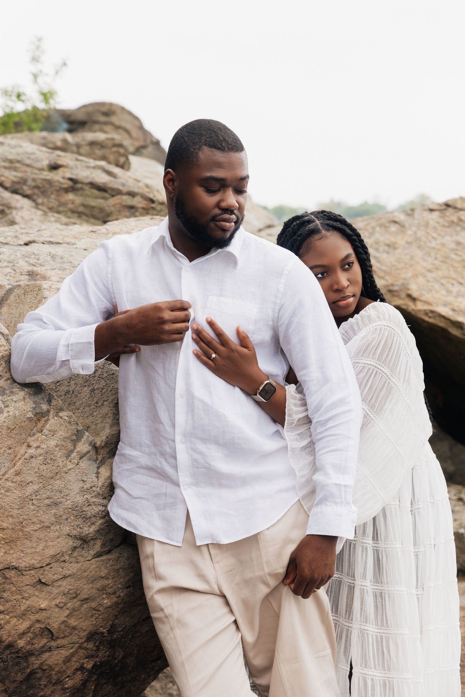 A Black couple in white outfits leans against large rocks outdoors, the woman resting her hand on the mans chest.