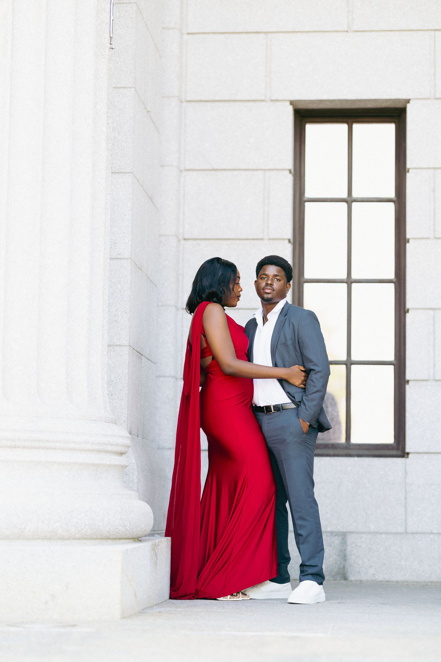 Elegant couple posing at a white stone building, woman in red cape gown, man in gray suit, romantic engagement photo.
