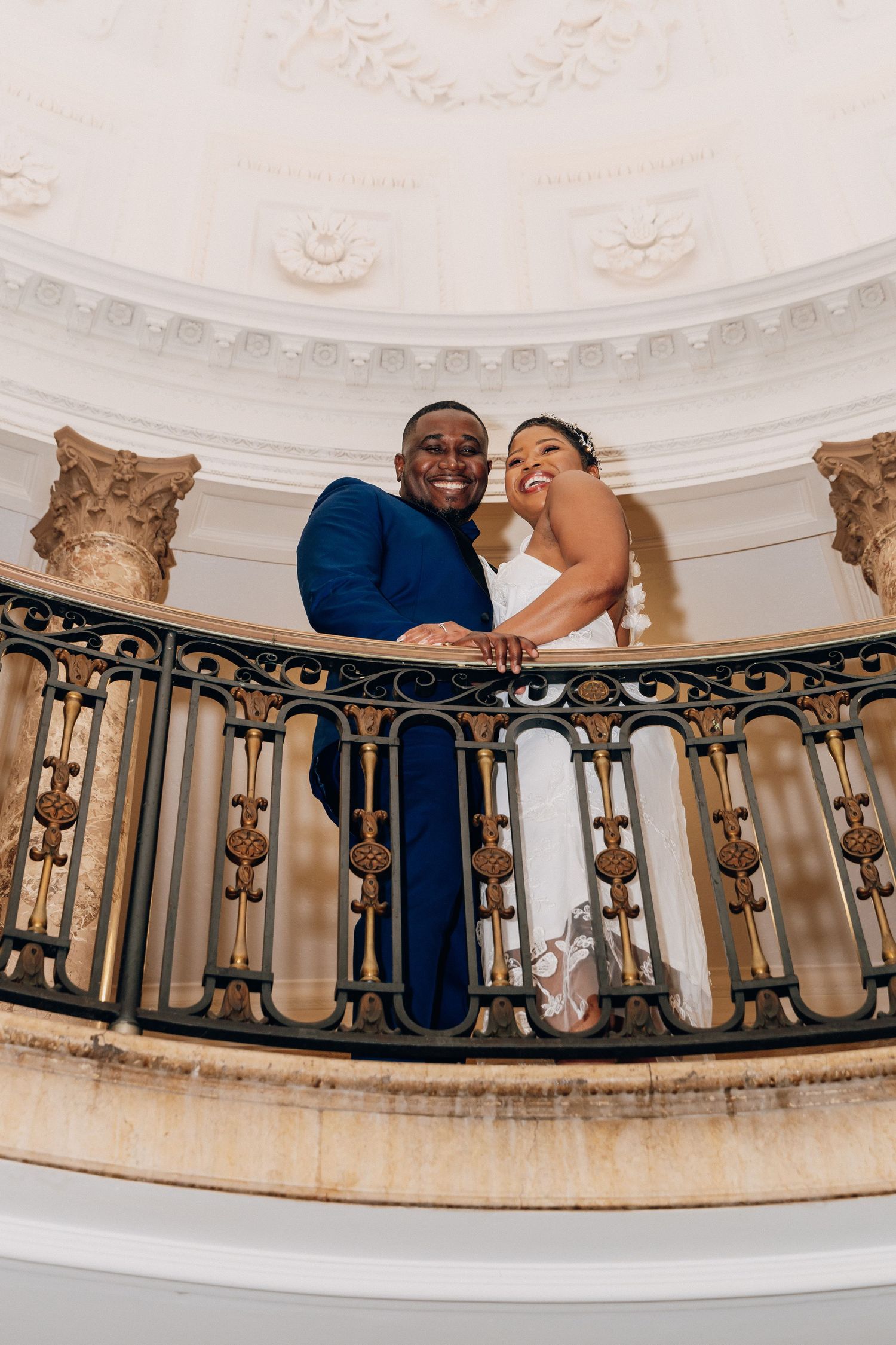 Smiling bride and groom lean on ornate iron balcony railing in elegant domed venue.