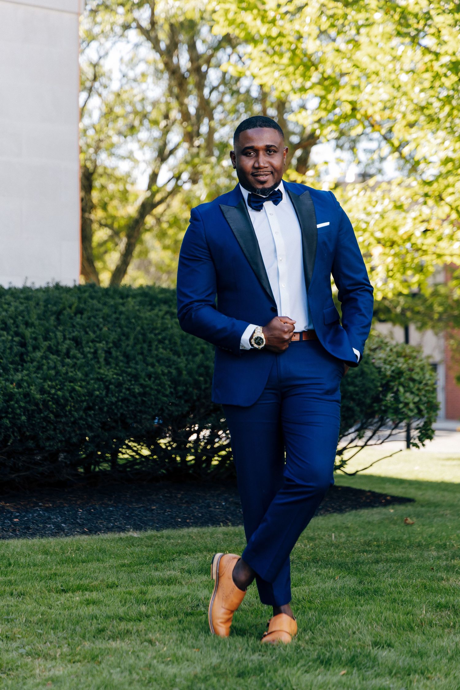 Confident groom in navy blue tuxedo with bow tie and tan shoes posing outdoors on green lawn.