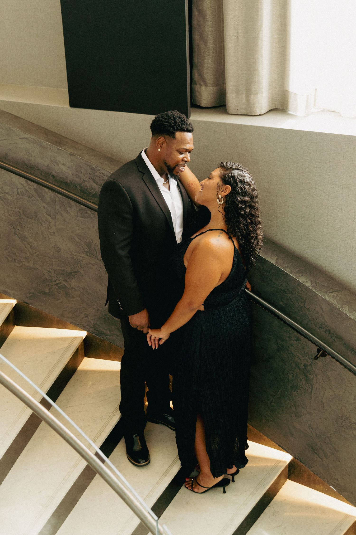 Couple in black formal wear share a romantic moment on elegant marble stairs in a modern venue.
