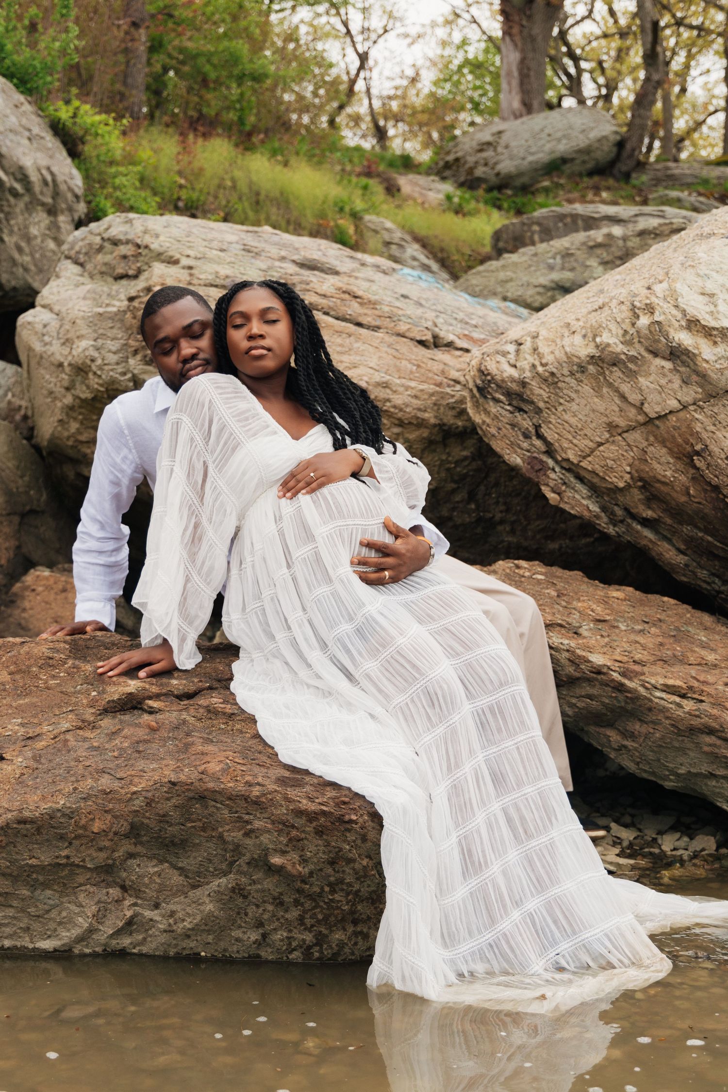 Pregnant Black couple in white outfits sitting on rocks near water, man embracing woman from behind in nature.