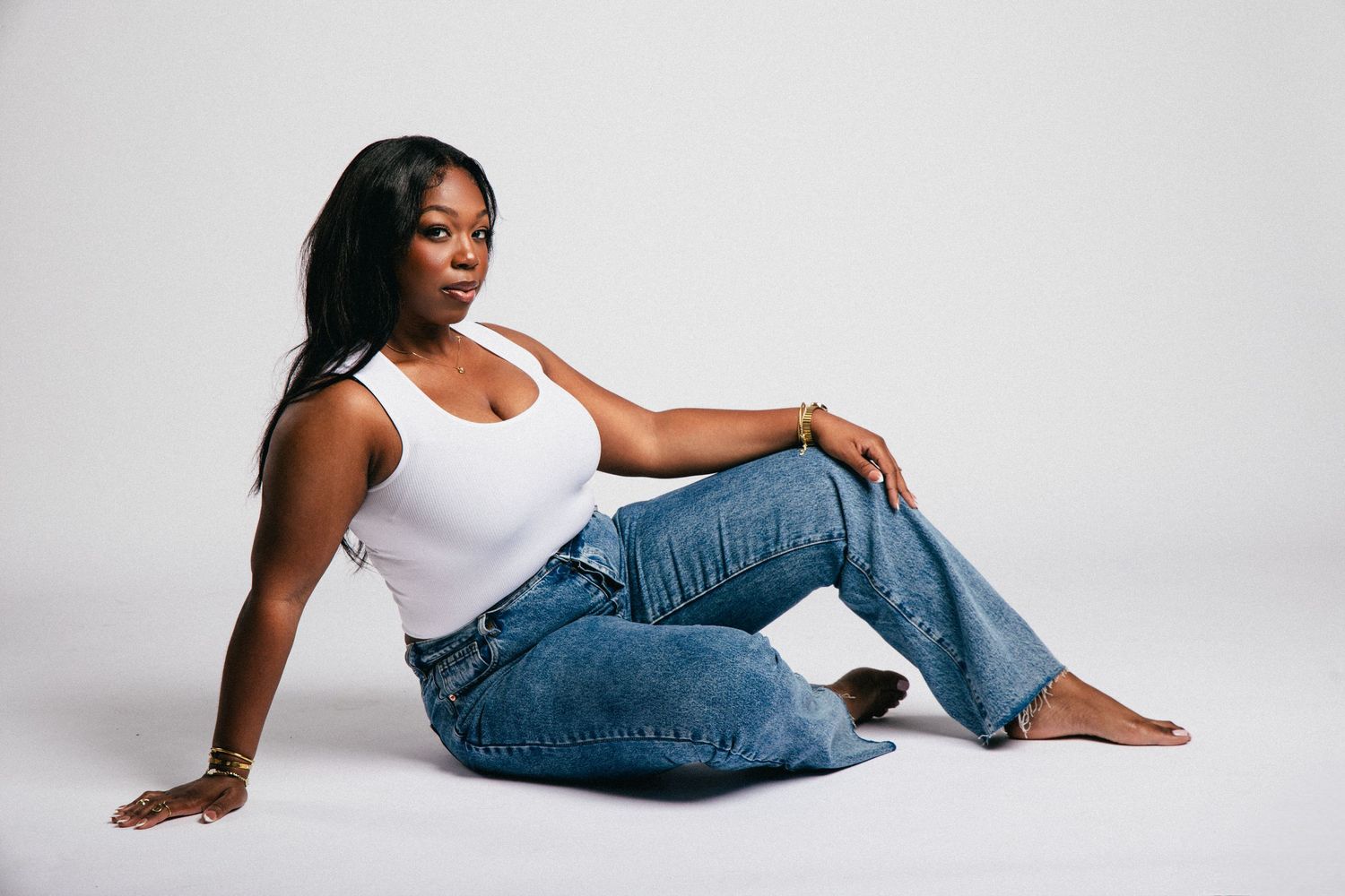 A Black woman sits on a white studio floor wearing a white tank top and blue jeans, posing confidently.