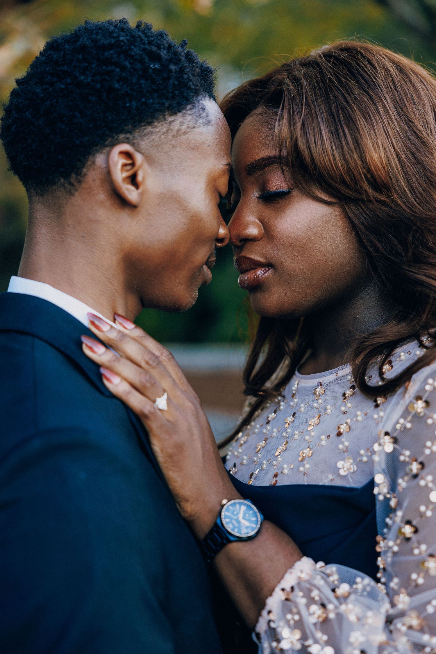 A romantic couple in elegant attire share an intimate moment, touching foreheads tenderly outdoors.