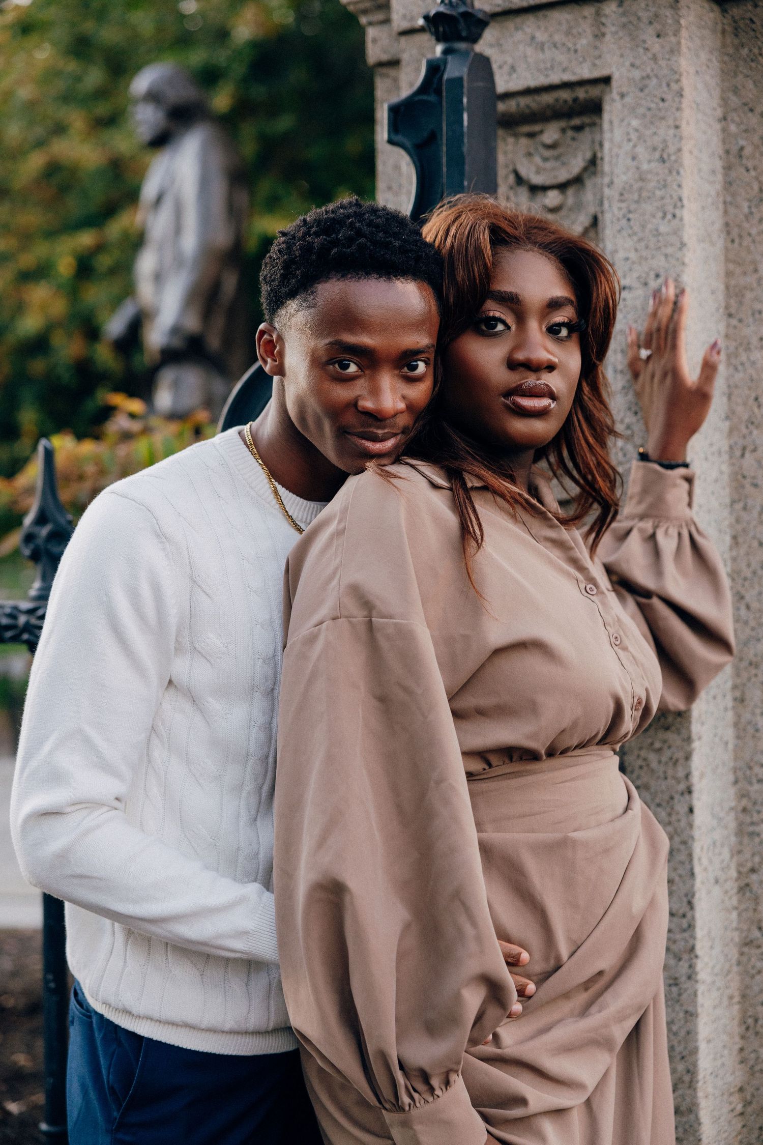 A young Black couple poses together outdoors near a stone monument, man in white sweater, woman in beige dress.
