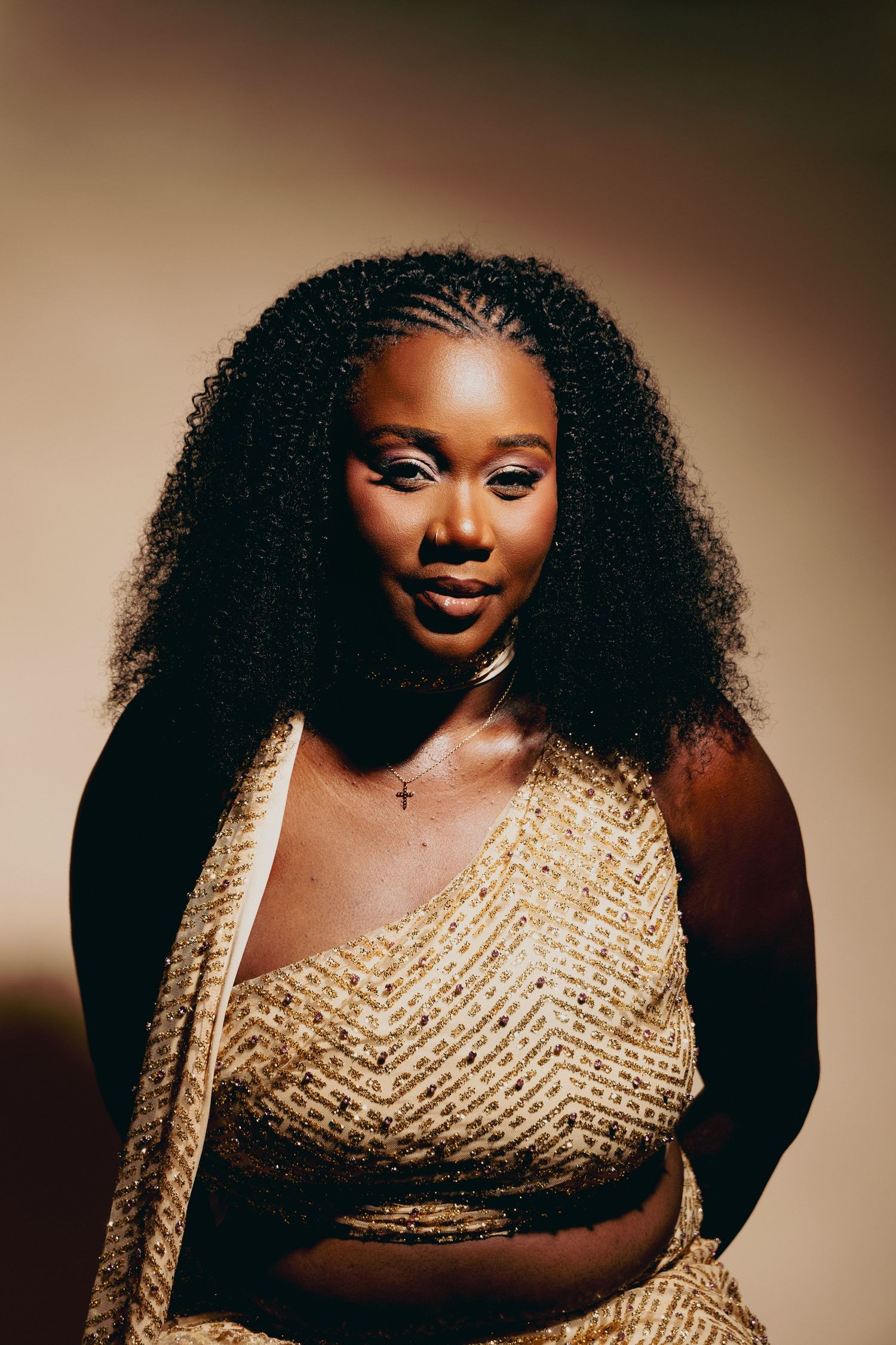 A confident Black woman in a gold sequined outfit with natural curly hair poses against a warm beige background.