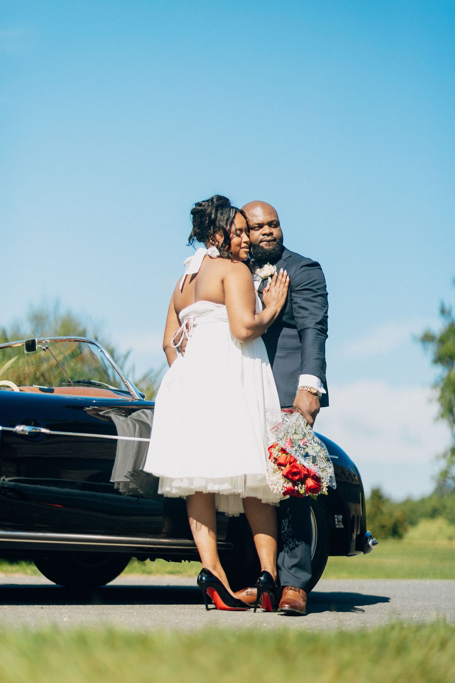 Black bride and groom embracing in front of a classic black vintage convertible under a bright blue sky.