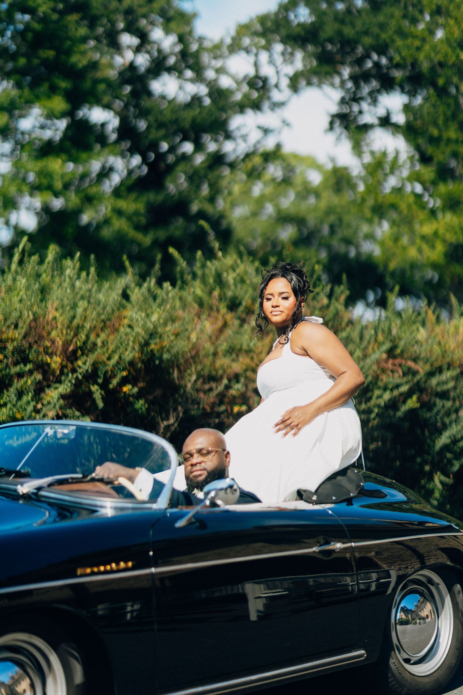 Bride in white halter dress sits on vintage black convertible hood while groom sits in drivers seat outdoors.