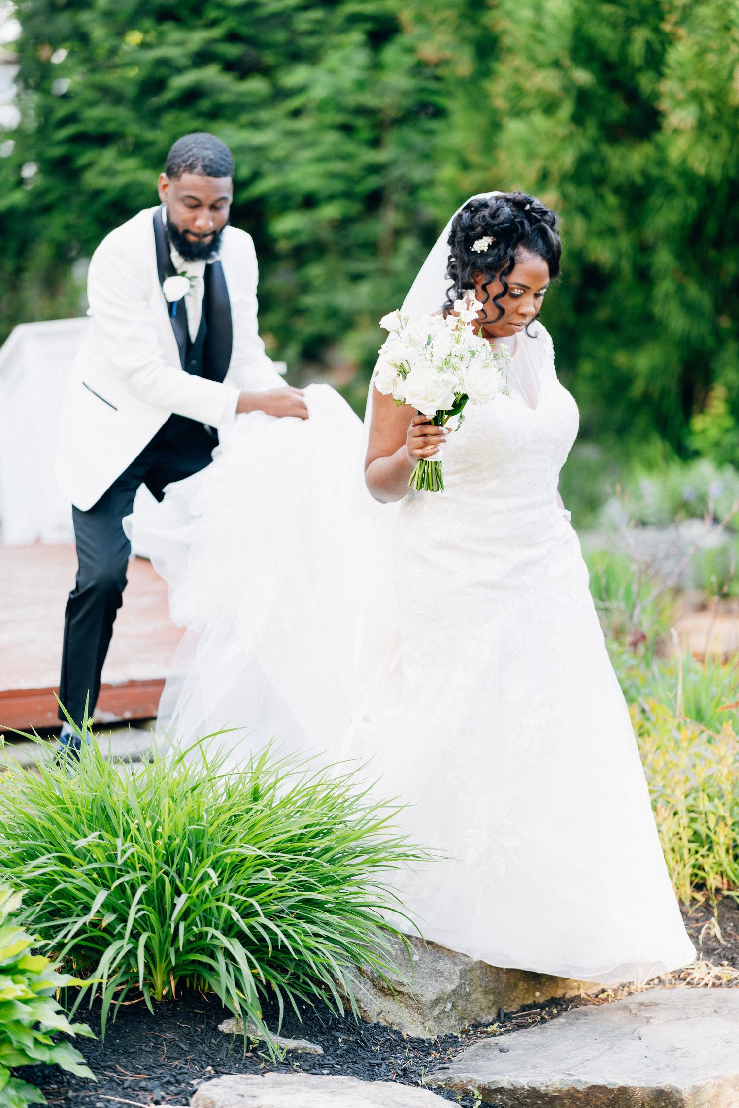 A groom in a white tuxedo helps his bride in a white gown navigate garden steps during their outdoor wedding.