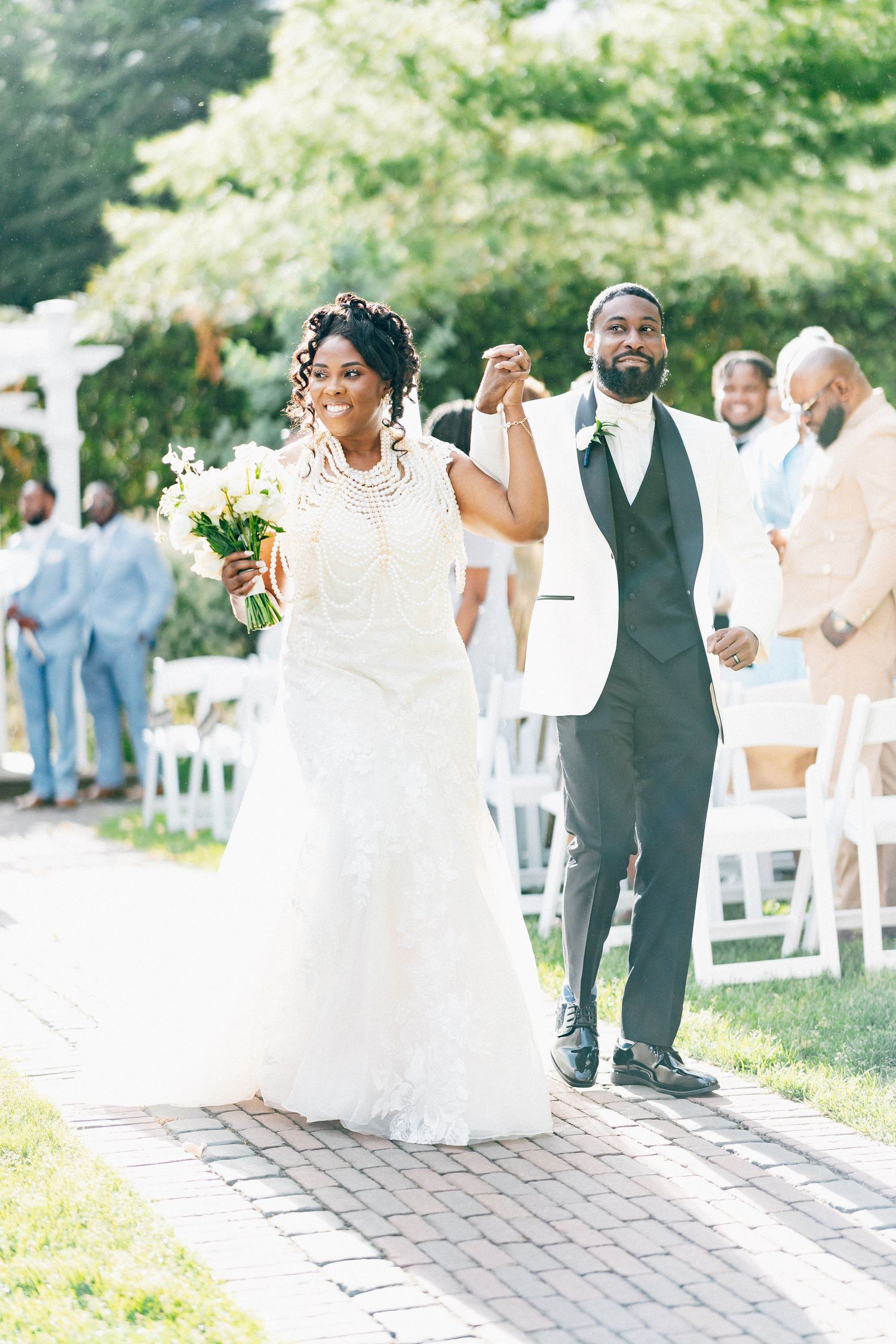 Joyful newlyweds walk down the aisle holding hands raised in celebration at an outdoor garden wedding ceremony.