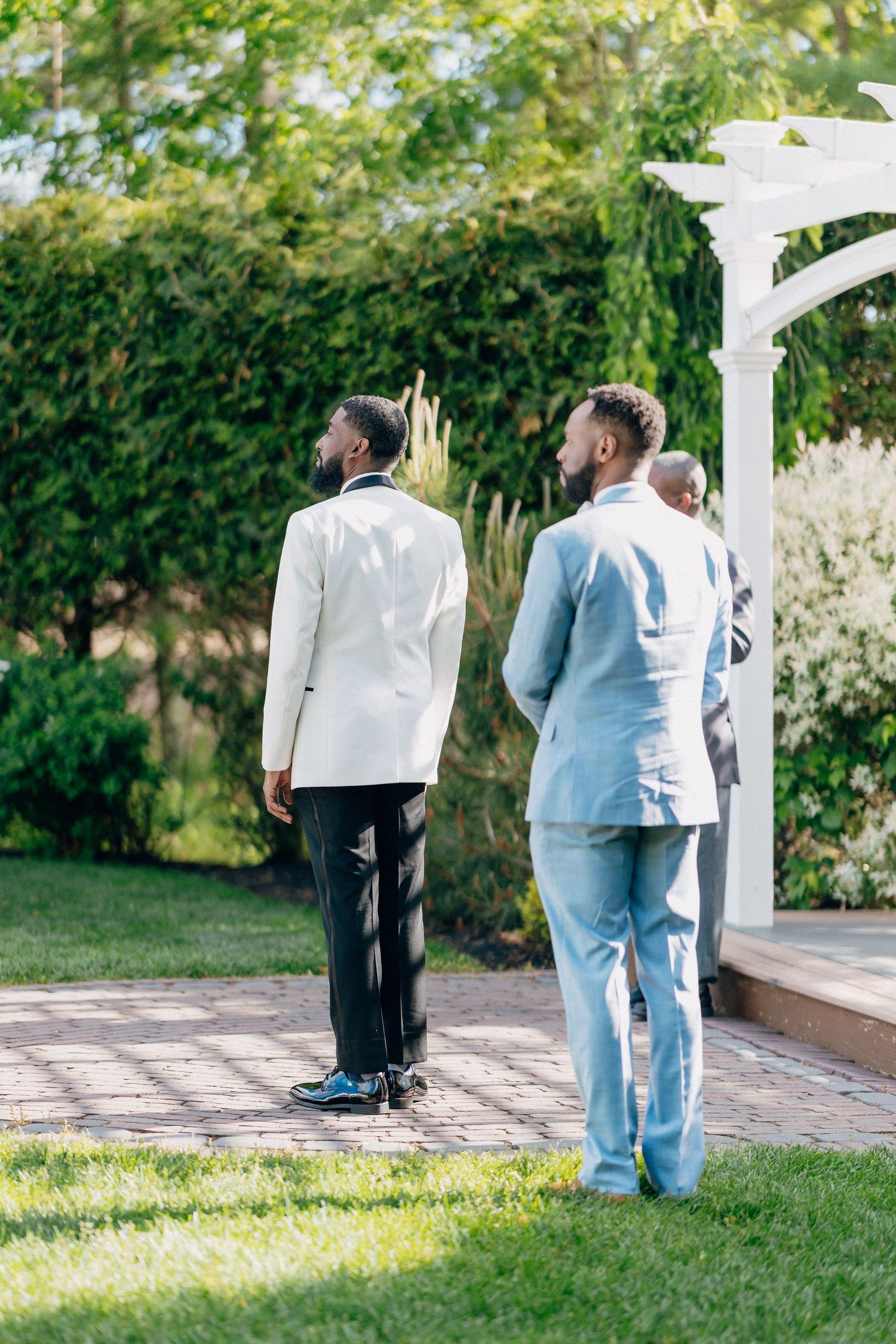 Groom in white jacket and groomsman in light blue suit standing at white wedding arch outdoors.