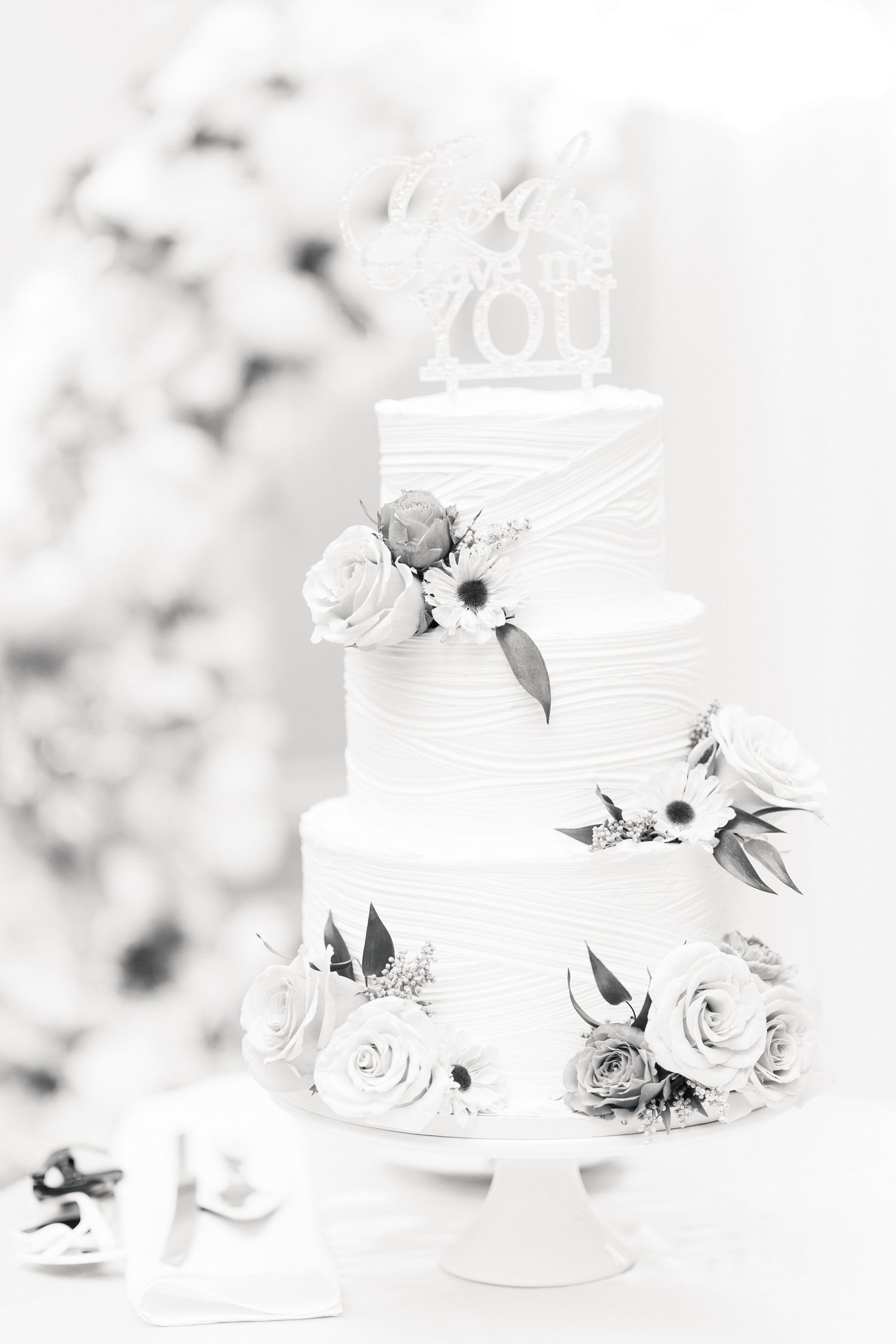 Elegant three-tier white wedding cake decorated with roses and floral topper in black and white.