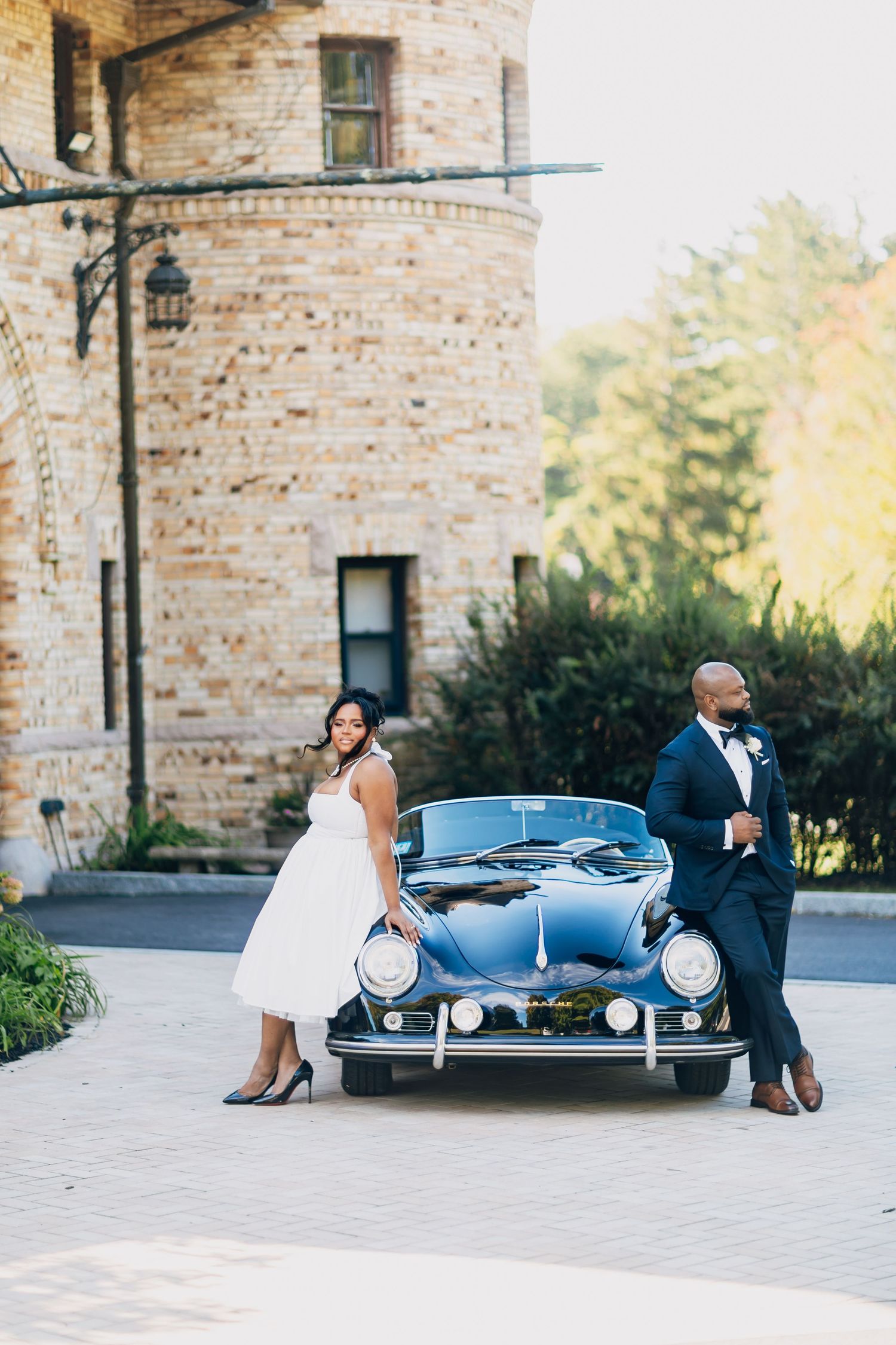 Bride and groom posing with a vintage black Porsche convertible in front of a stone castle venue.