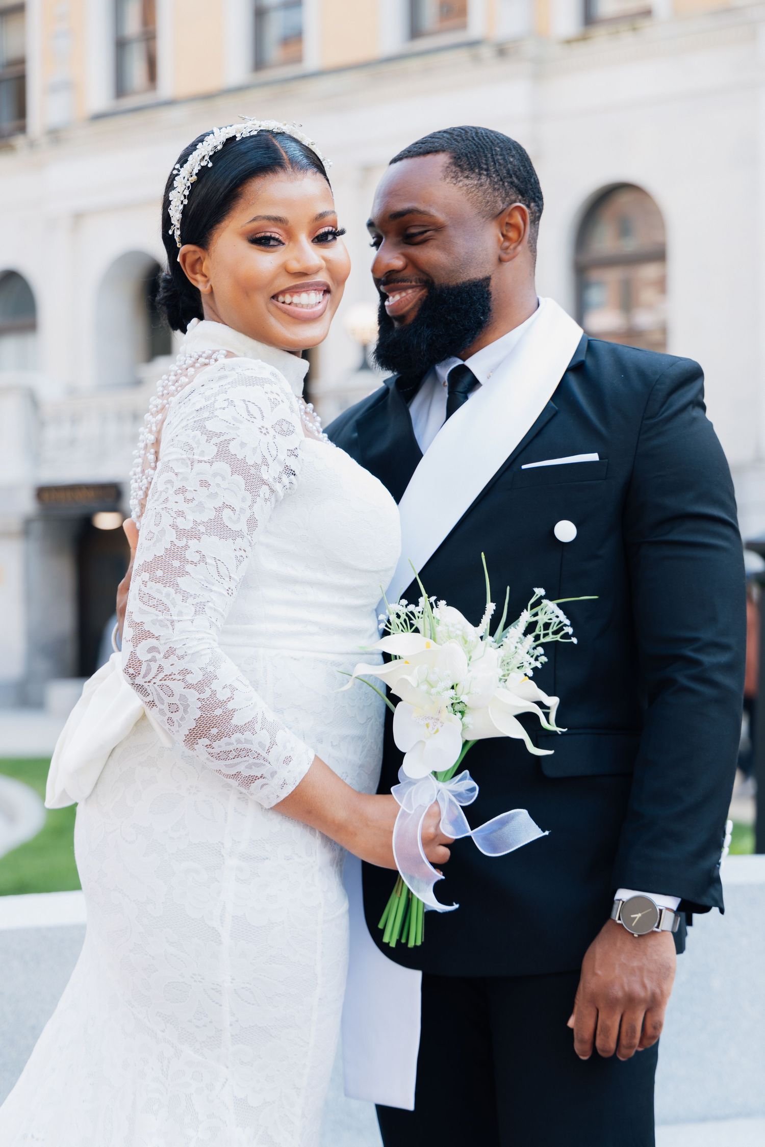 A smiling Black bride in lace gown holds white bouquet beside groom in black tuxedo outdoors.