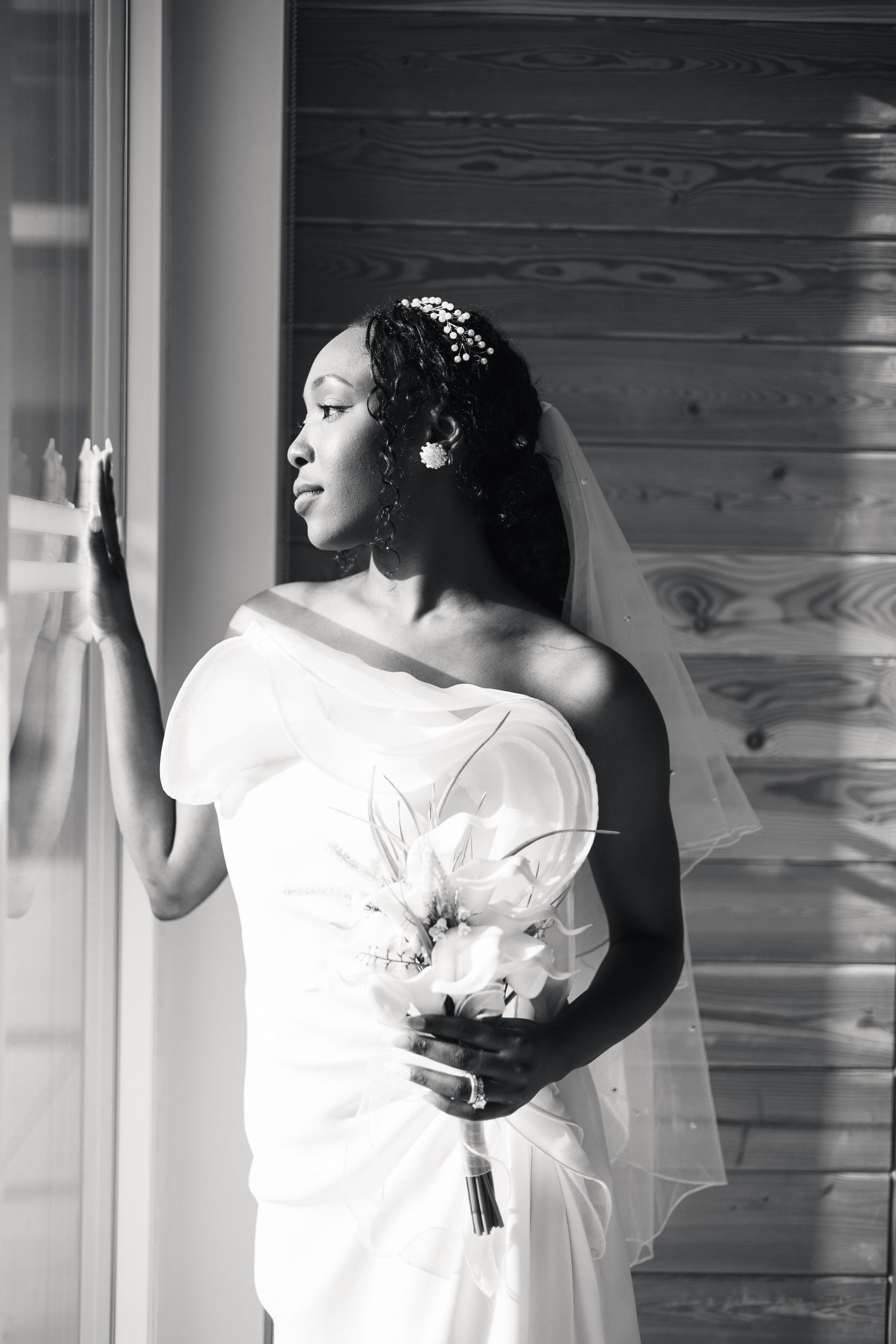A black and white portrait of a bride in a white gown and veil holding a bouquet by a sunlit window.