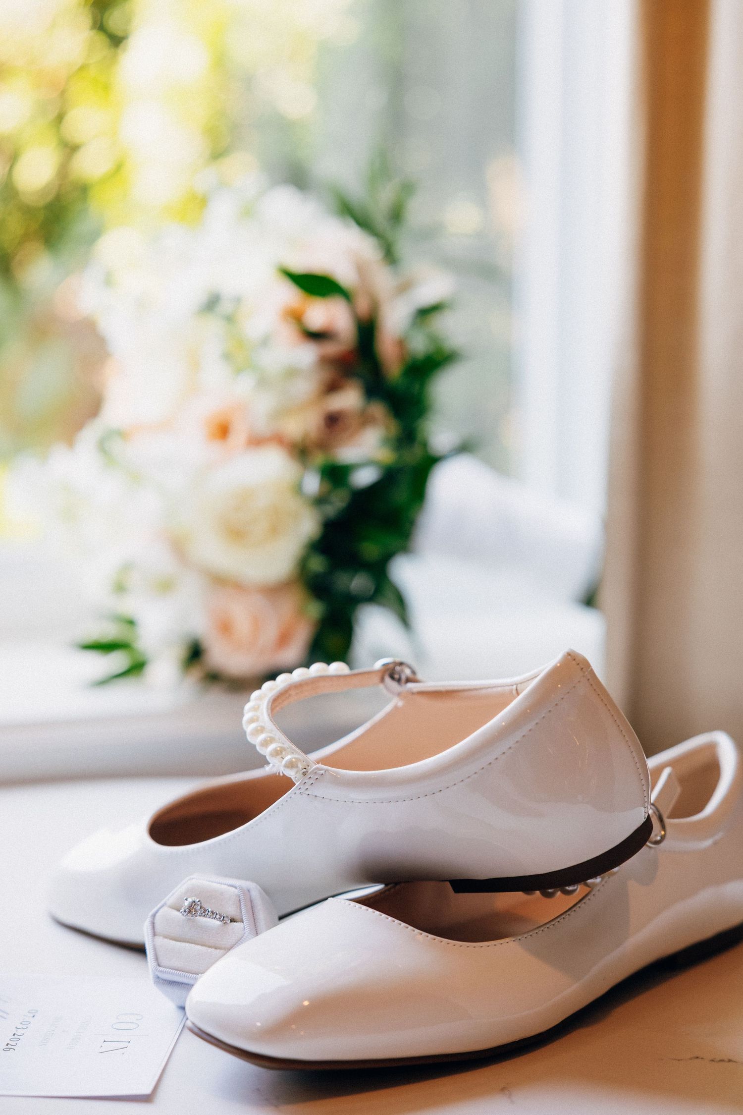 White patent leather bridal flats with pearl ankle strap beside wedding rings and bouquet of roses near window.