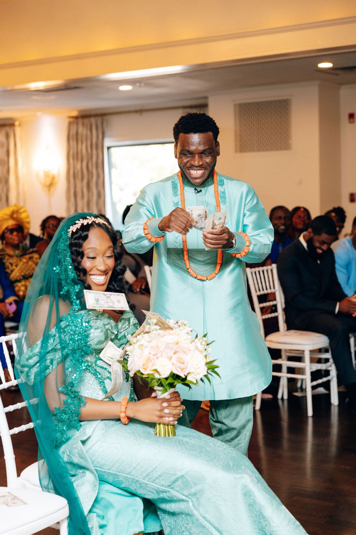 A laughing groom in a teal African suit stands beside his smiling bride holding a white bouquet.