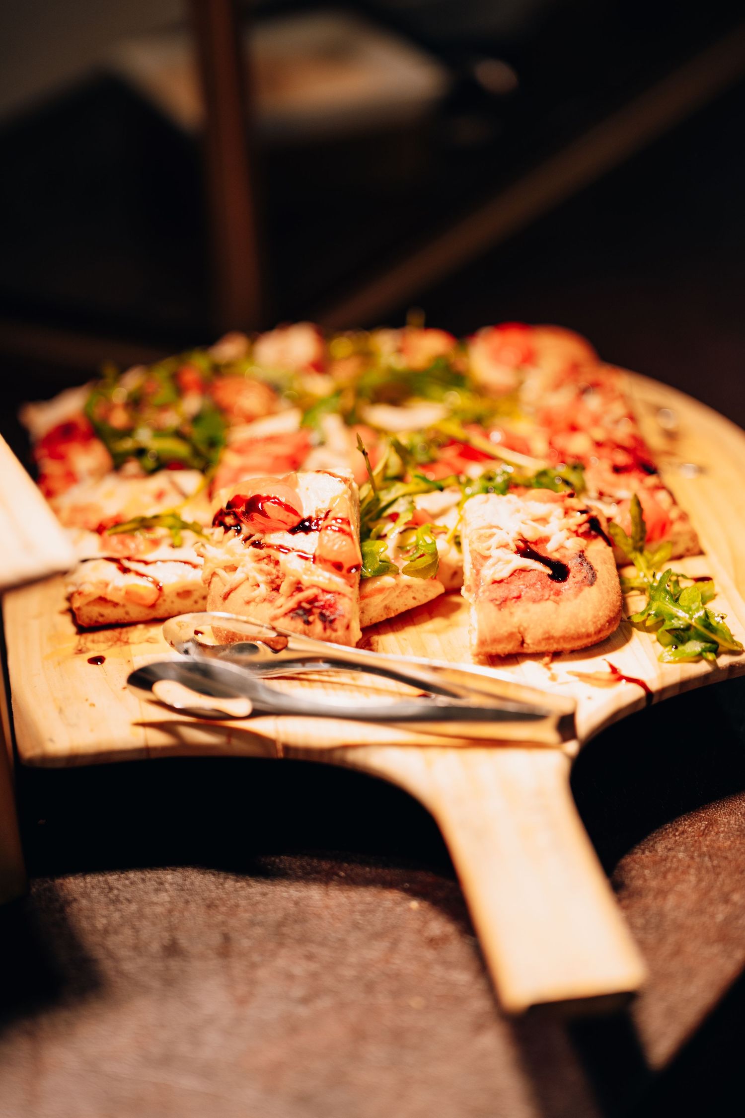 Sliced gourmet pizza with fresh arugula and toppings served on a wooden paddle board at an event.