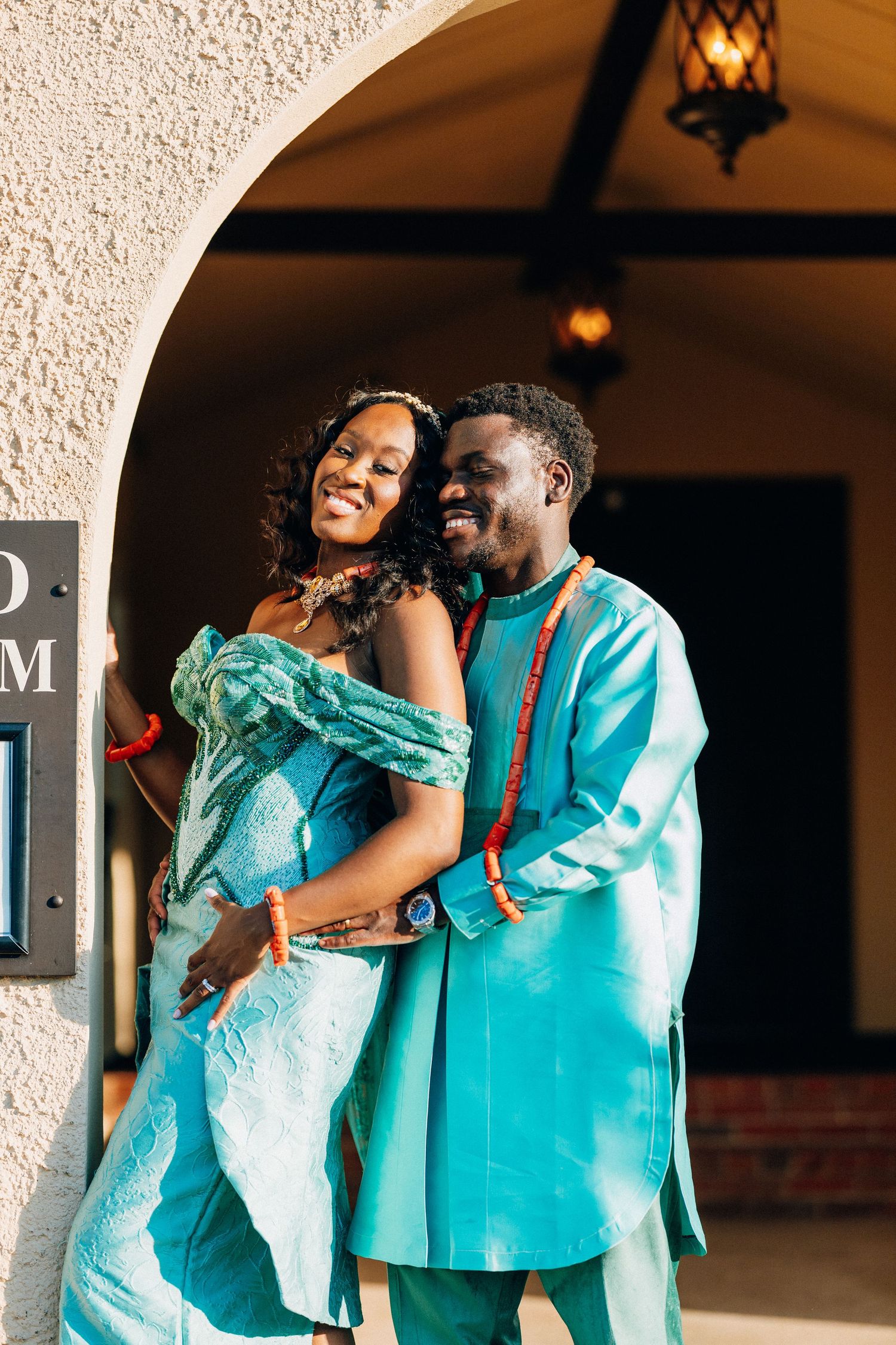 Couple in matching teal African attire embrace under archway.