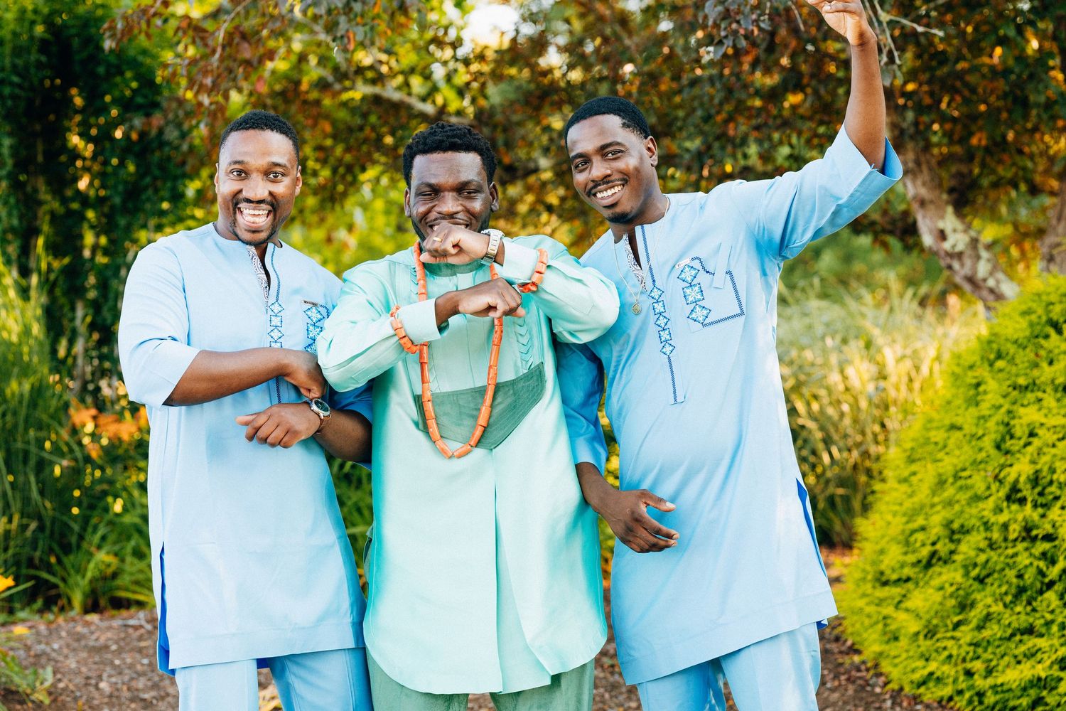 Three men in matching light blue African traditional attire smiling and taking a selfie outdoors.
