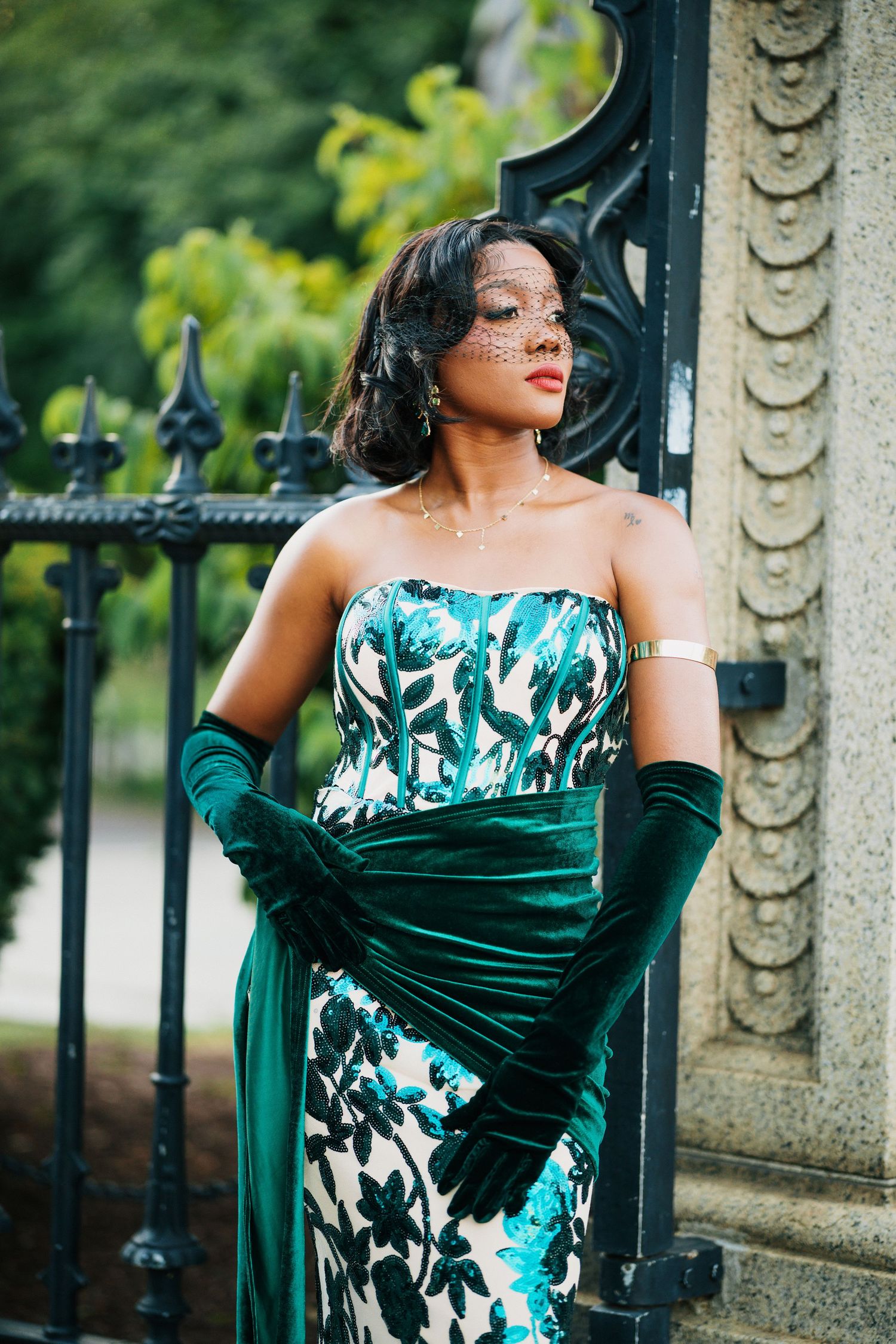 Elegant Black woman in green floral strapless gown with velvet gloves and veil poses by ornate iron gate.