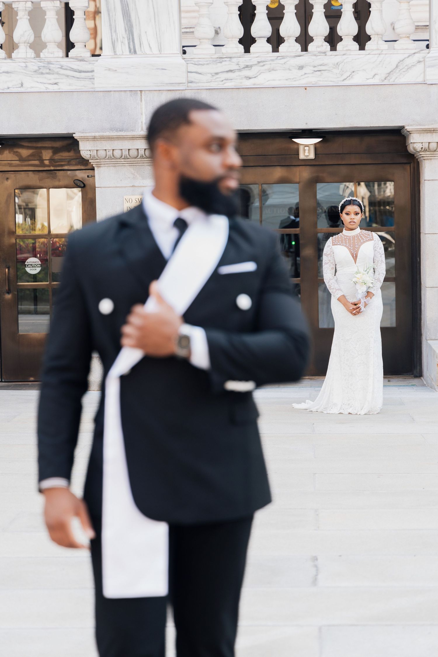 A groom in a black tuxedo stands in the foreground as his bride in a white gown poses behind him outdoors.