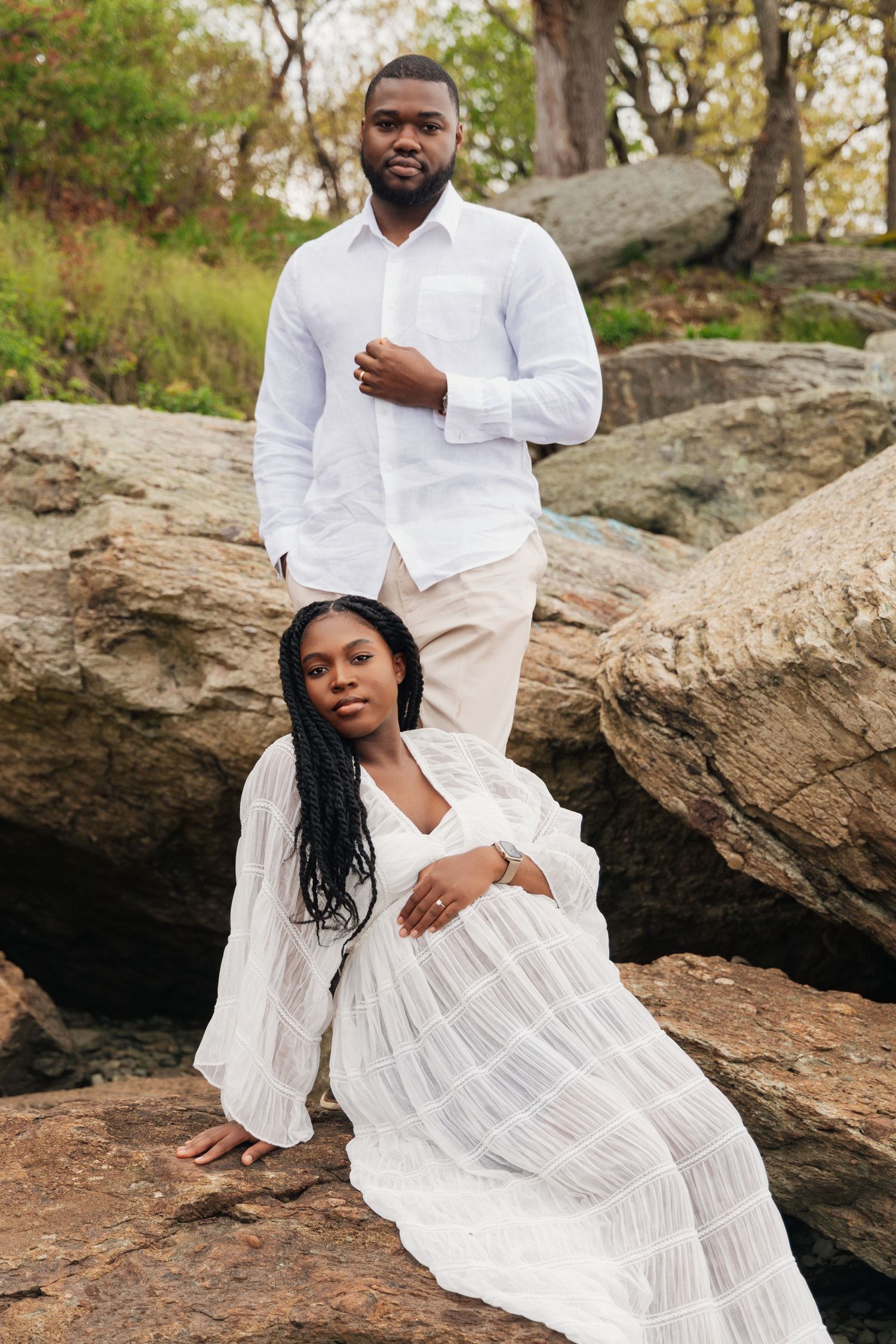 A couple in matching white outfits poses on rocky terrain, woman in flowing dress seated while man stands behind her.
