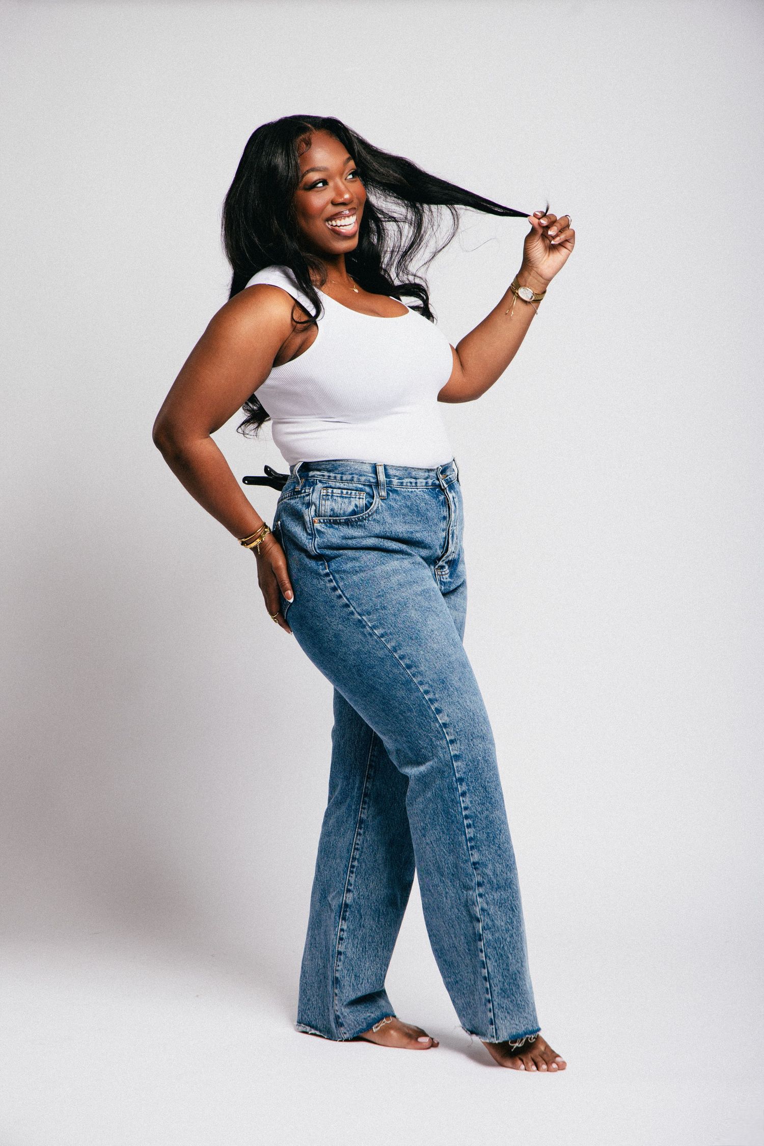 Smiling Black woman in white tank top and wide-leg blue jeans, posing playfully while holding her long hair.