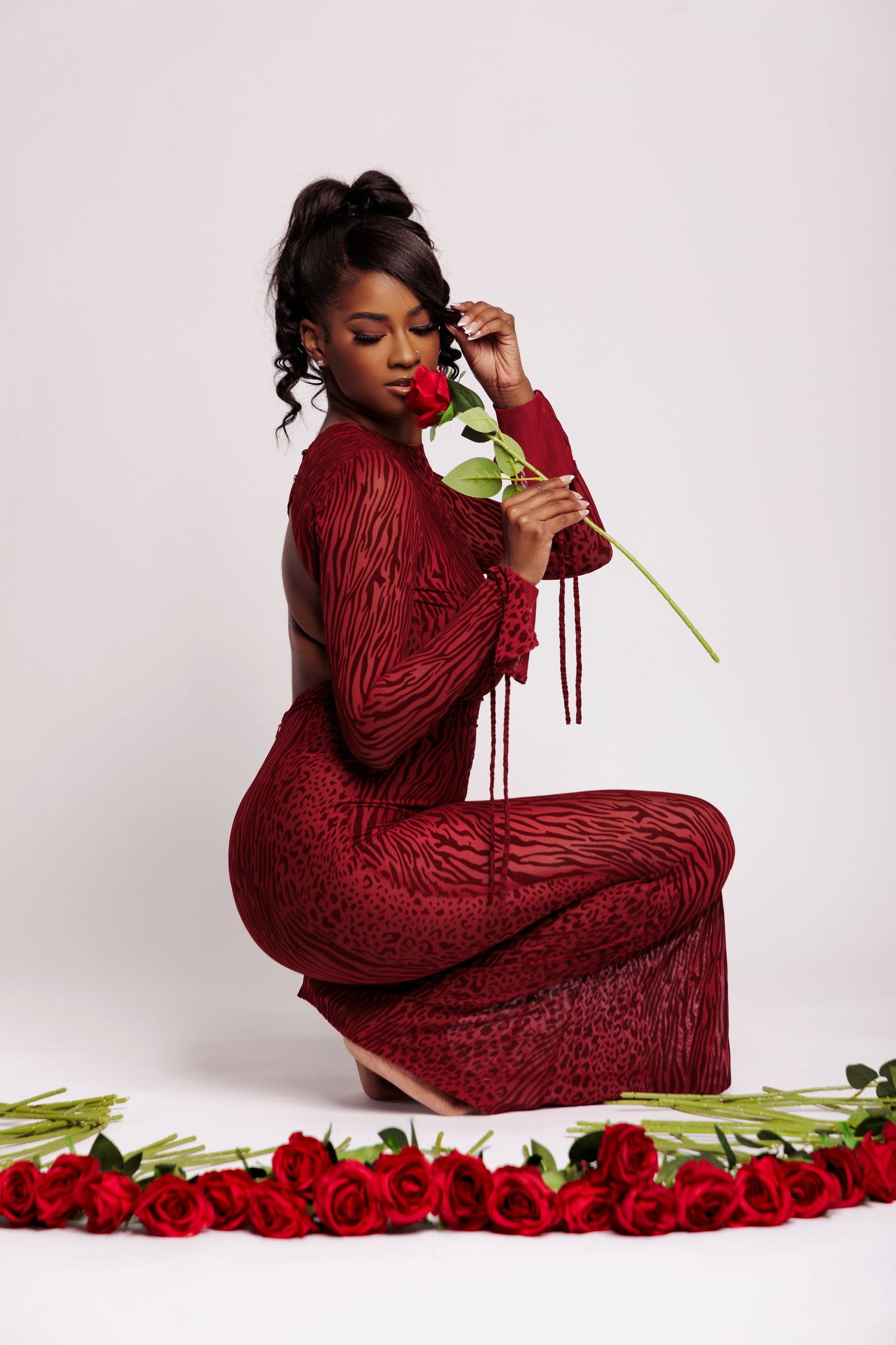 A Black woman in a red sequin gown kneels gracefully, smelling a red rose surrounded by roses on white background.