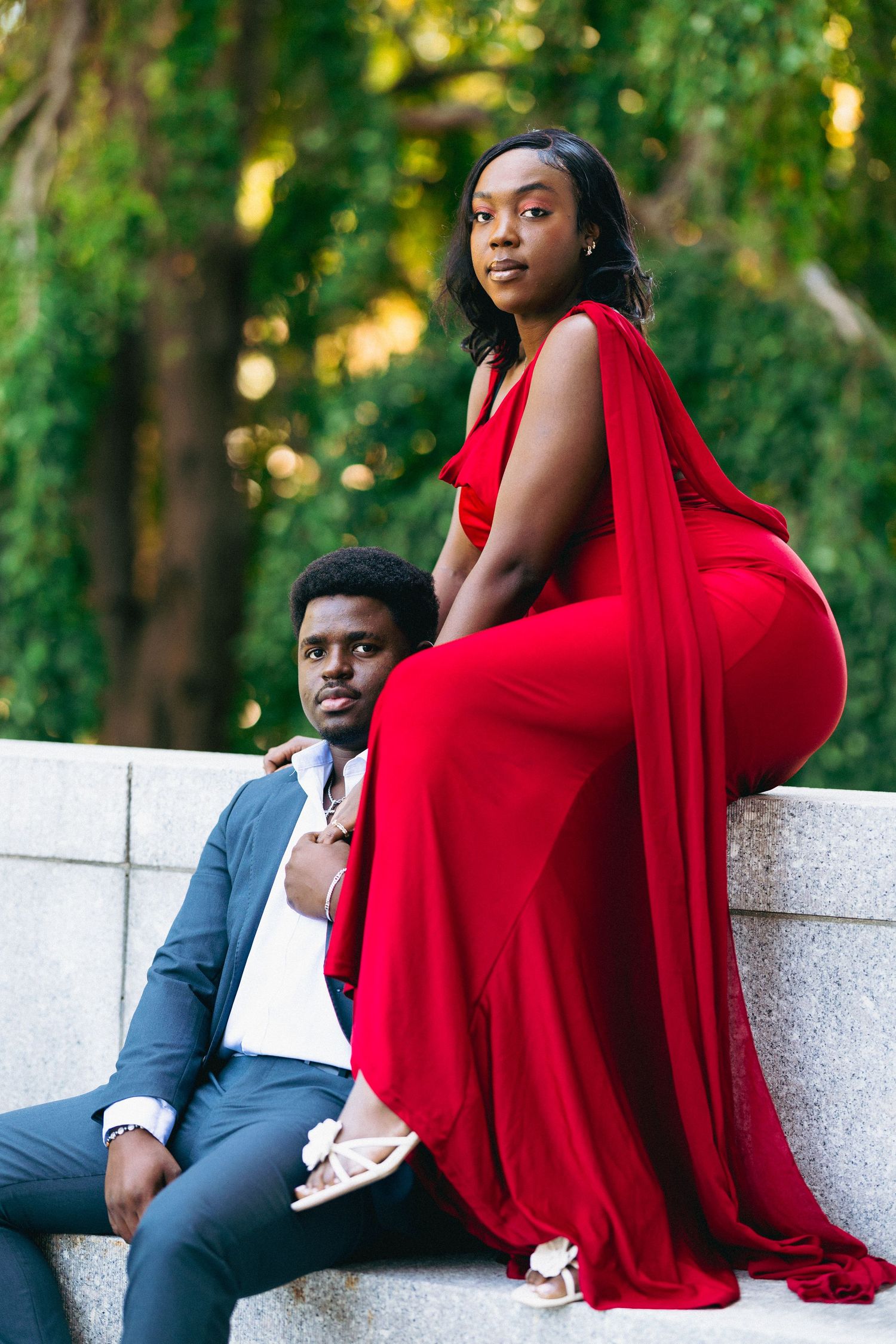A Black couple poses outdoors, woman in elegant red gown seated on stone wall, man in gray suit beside her.