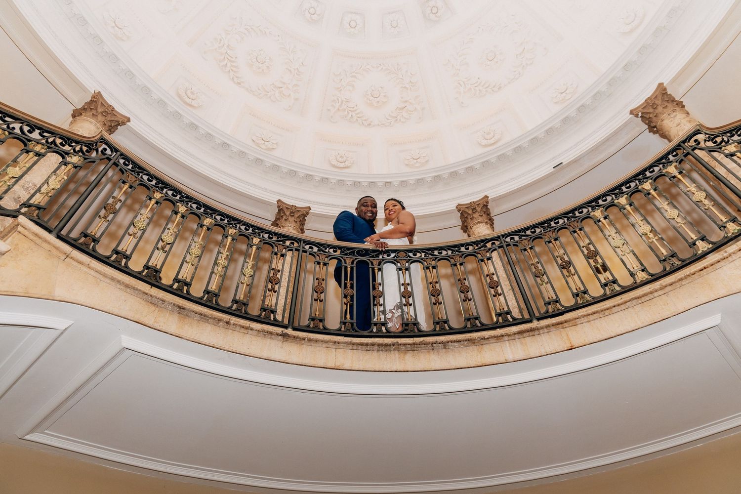 Bride and groom share a romantic moment on an ornate balcony beneath an elegant domed ceiling.