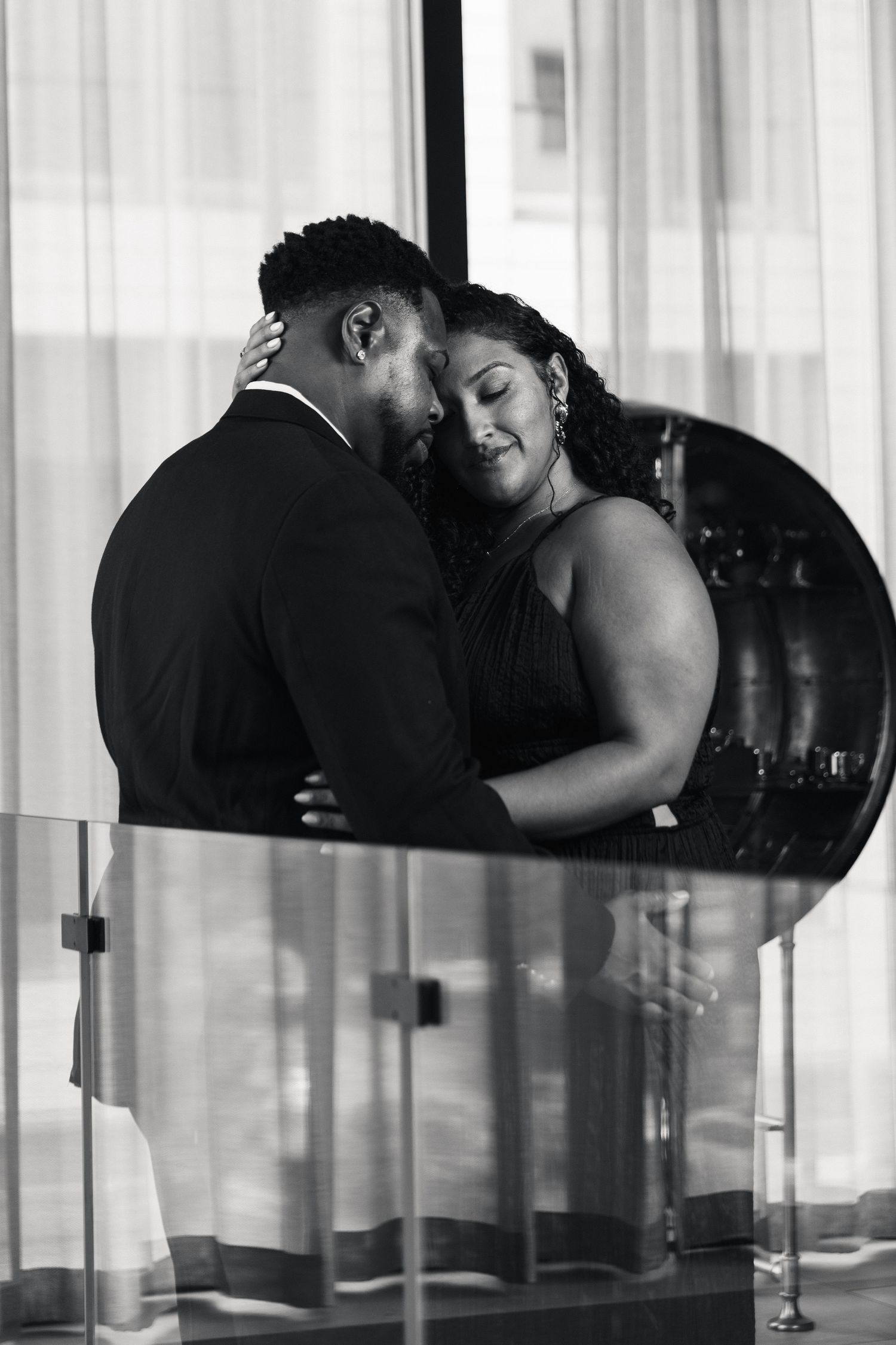 Black and white photo of a couple embracing tenderly, dressed in formal attire, sharing an intimate moment indoors.
