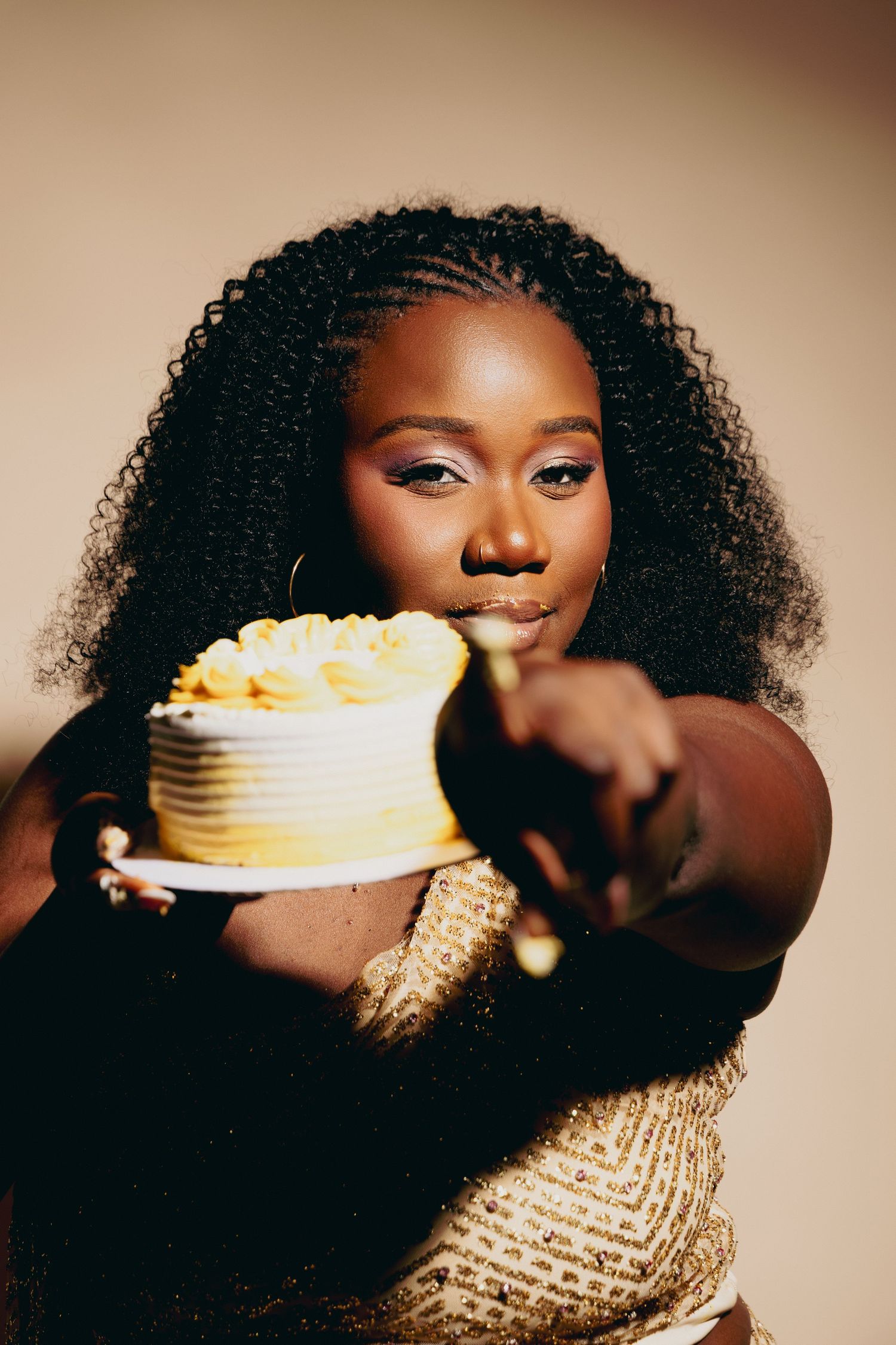 A young Black woman with curly hair offers a yellow ombre cake toward the camera while wearing a gold sequin outfit.