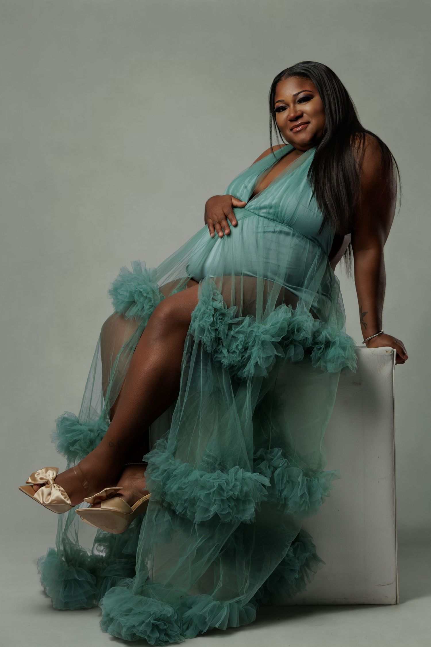 Pregnant woman in teal ruffle tulle maternity gown sitting gracefully during professional studio photo shoot.