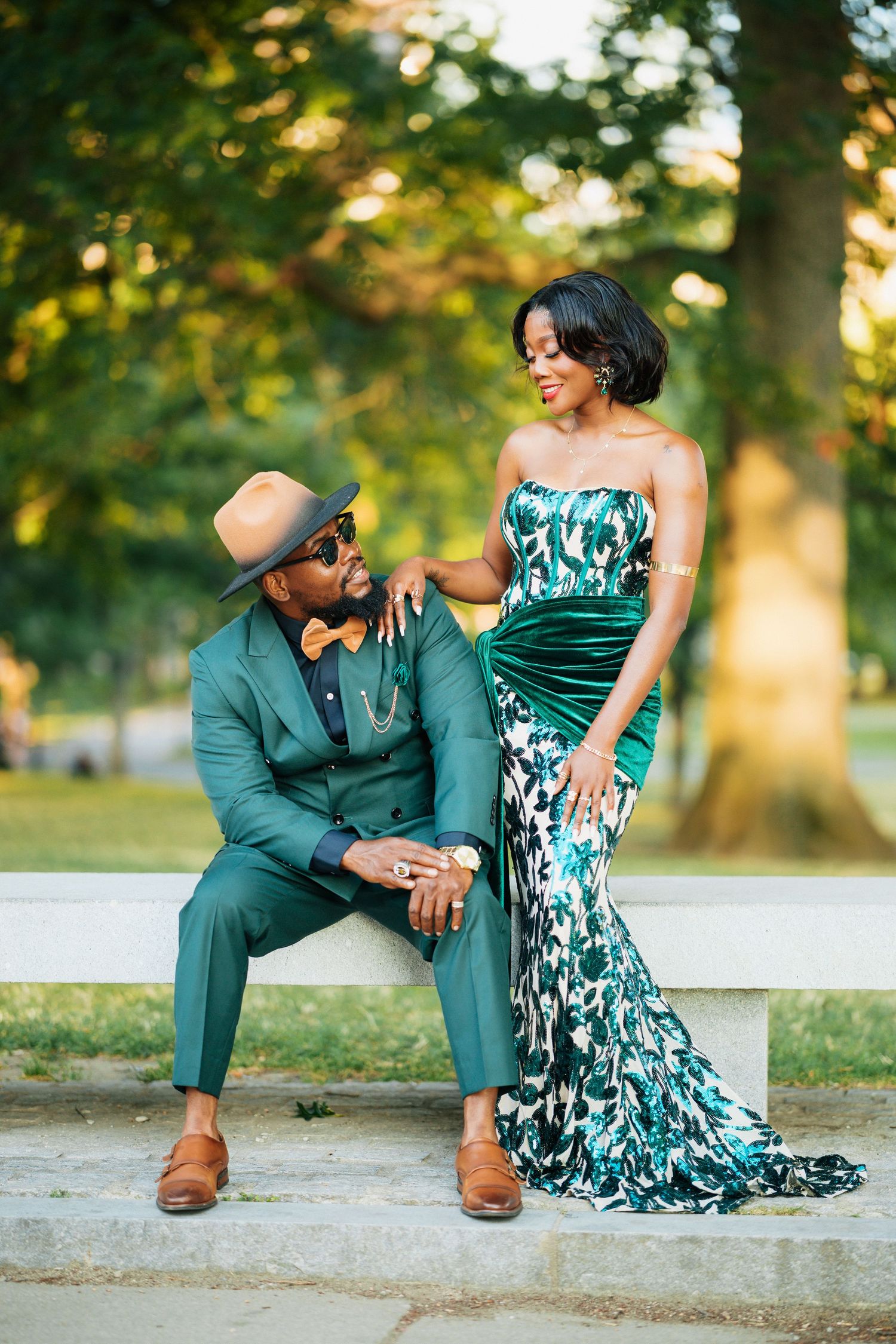 Stylish Black couple in matching emerald green outfits posing on stone bench in park.