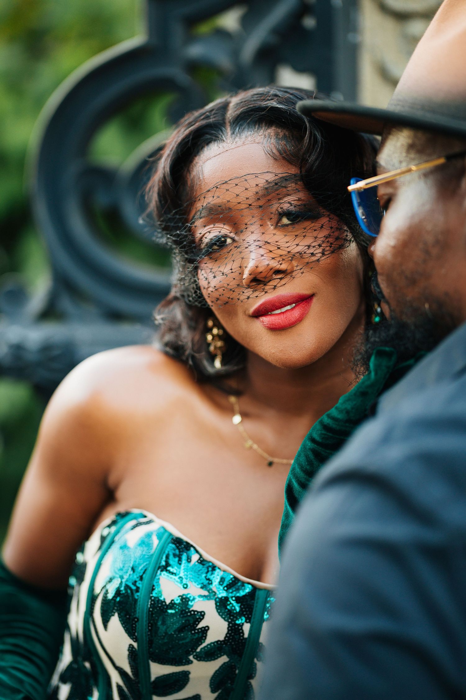 Elegant woman in green sequin dress with veil fascinator poses with a man in a hat outdoors.