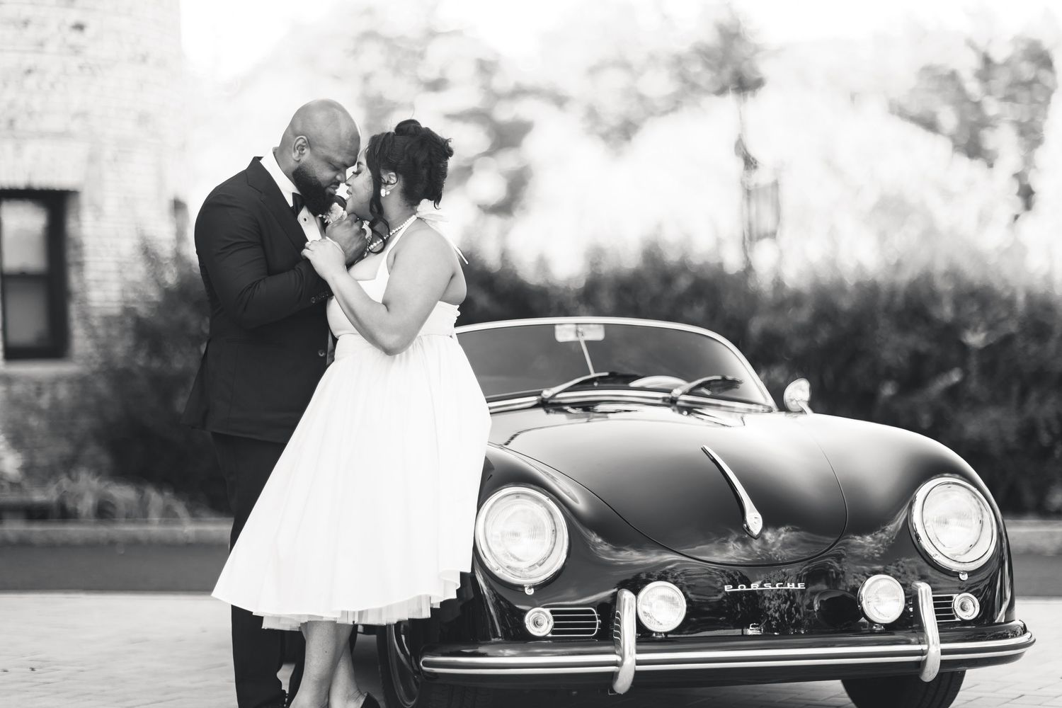 Black and white wedding photo of couple kissing beside a vintage classic convertible sports car outdoors.