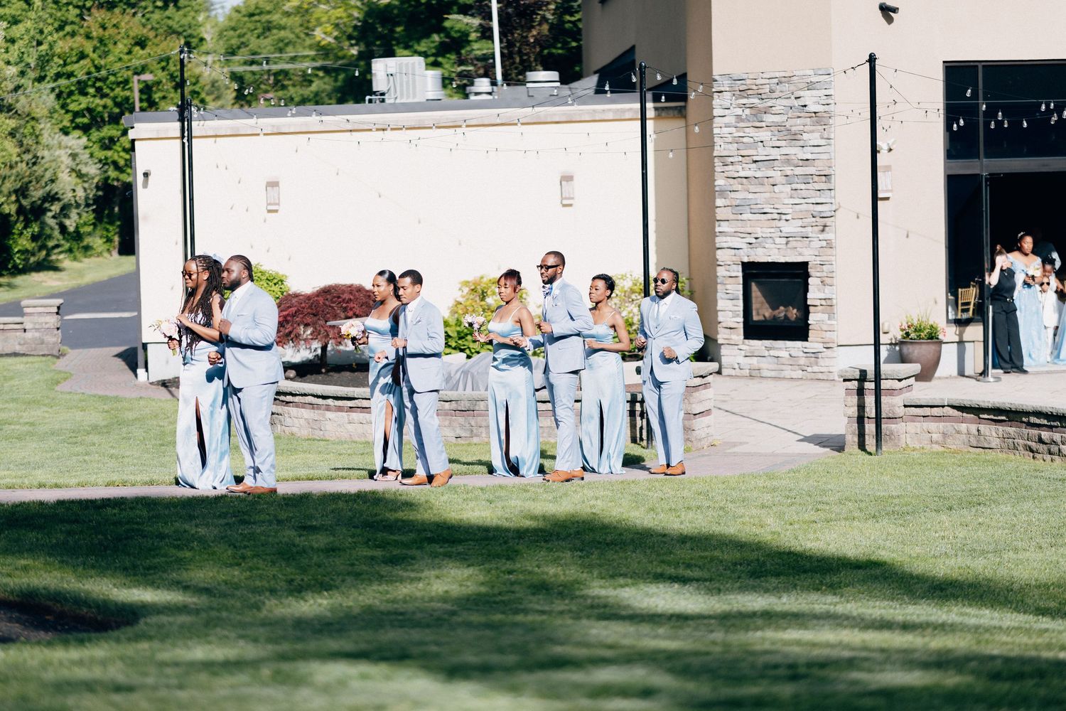 Wedding party in light blue attire gathered on green lawn outside stone venue on sunny day.
