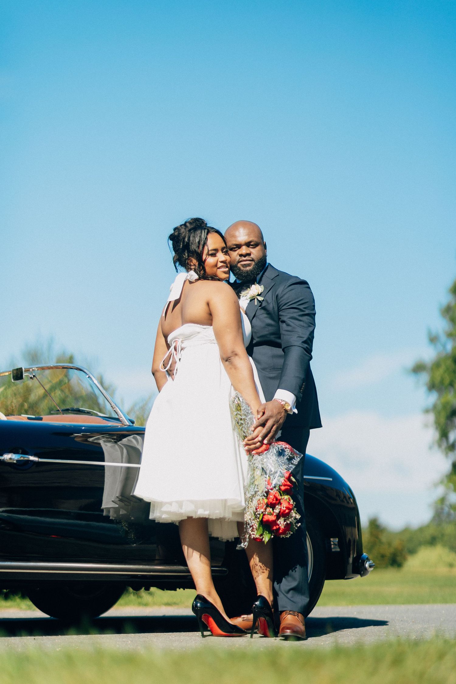 Wedding couple embraces beside classic black convertible car under bright blue sky on sunny day.