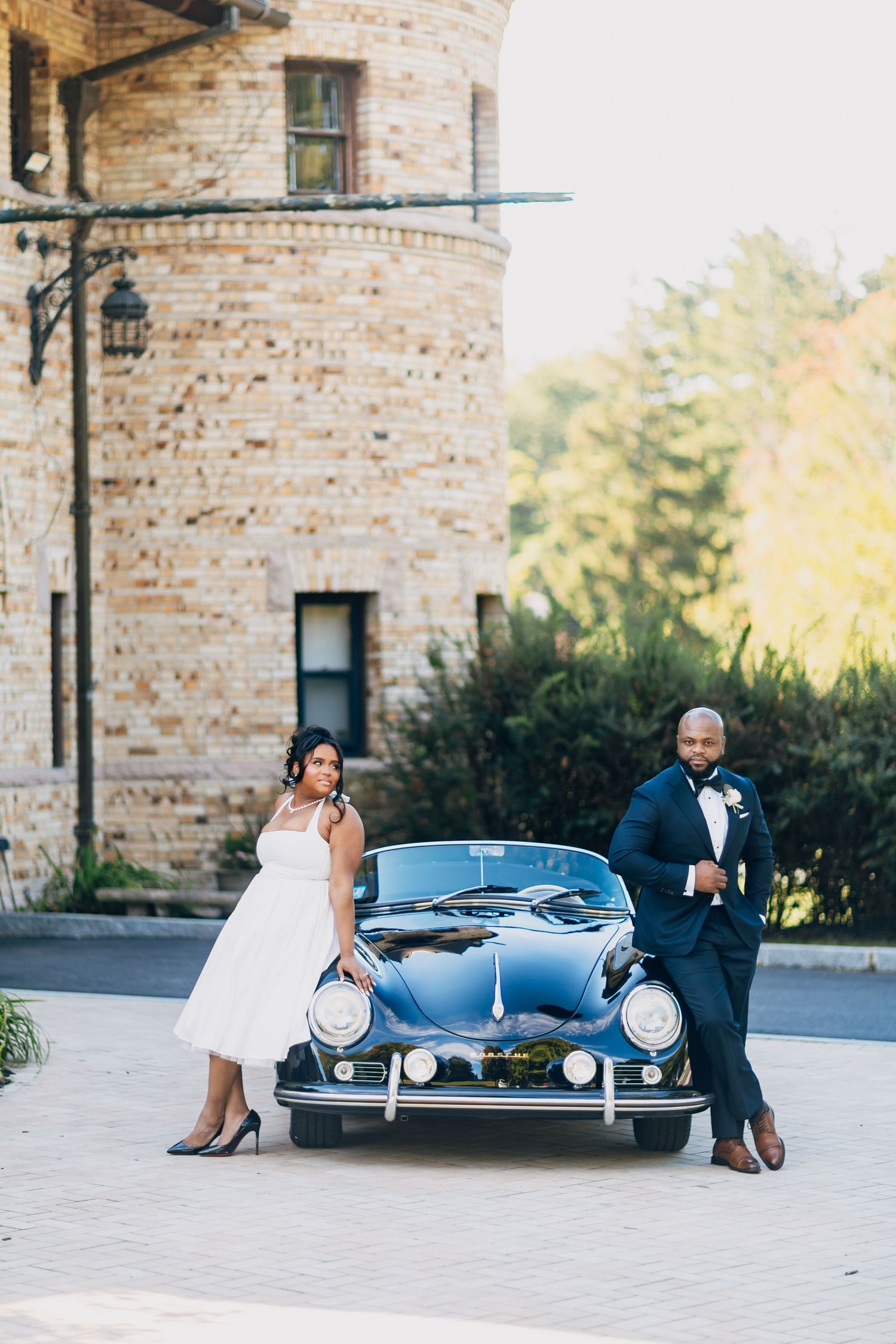 Bride and groom posing elegantly beside a vintage black Porsche 356 convertible in front of a stone building.