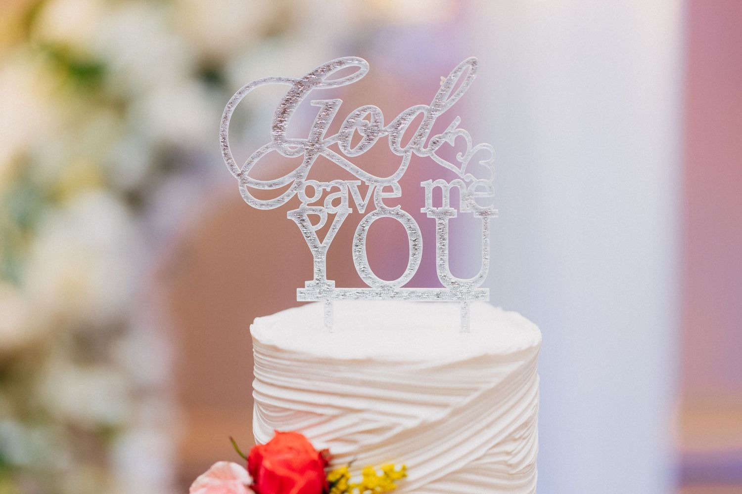 White wedding cake with silver glitter God Gave Me You cake topper, decorated with colorful flowers at the base.