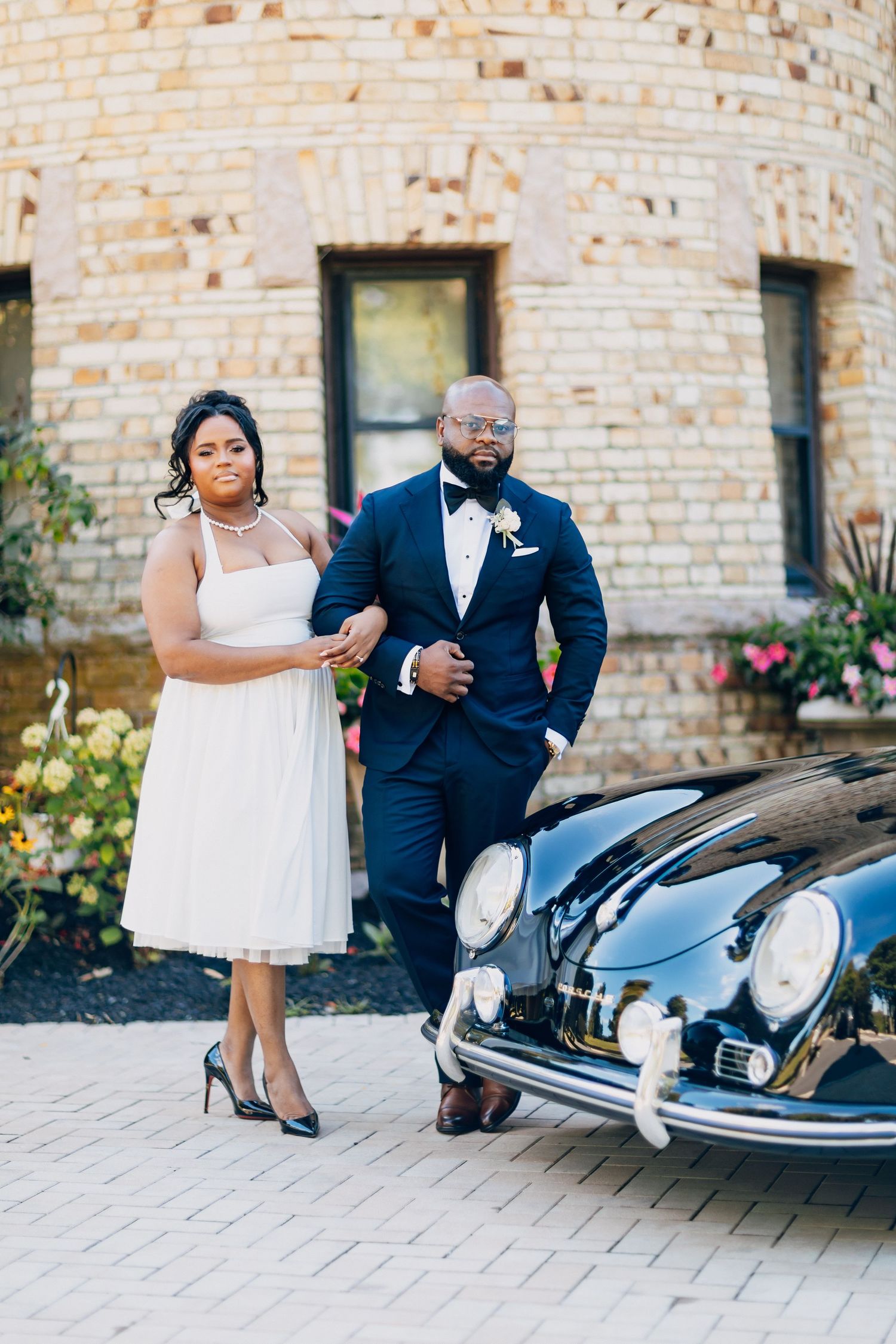 Bride in white dress and groom in navy tuxedo pose beside a classic black vintage car outside a brick building.